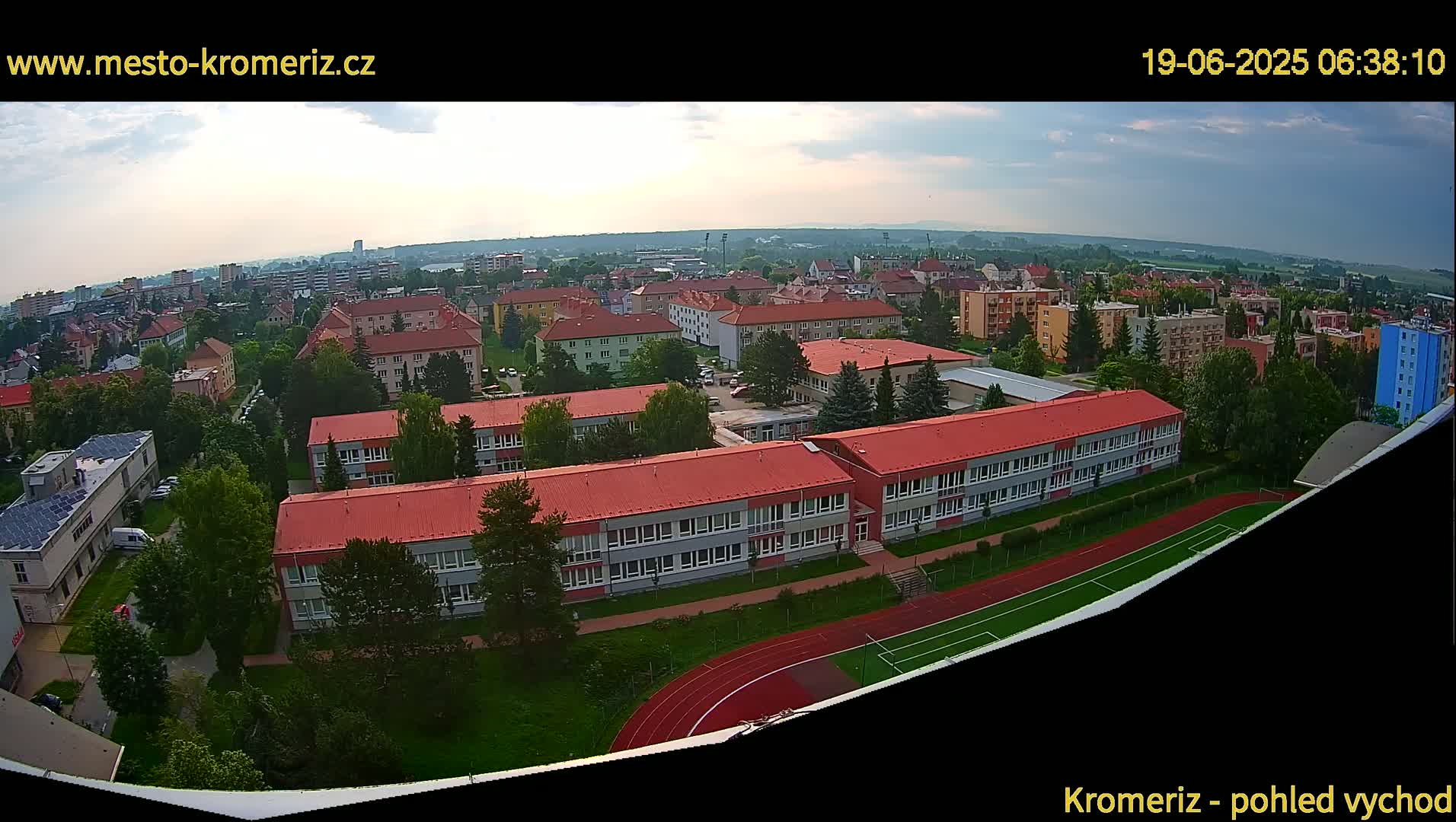 An aerial view shows a town under partly cloudy skies, focusing on a large school building with red roofs and a surrounding athletic field.