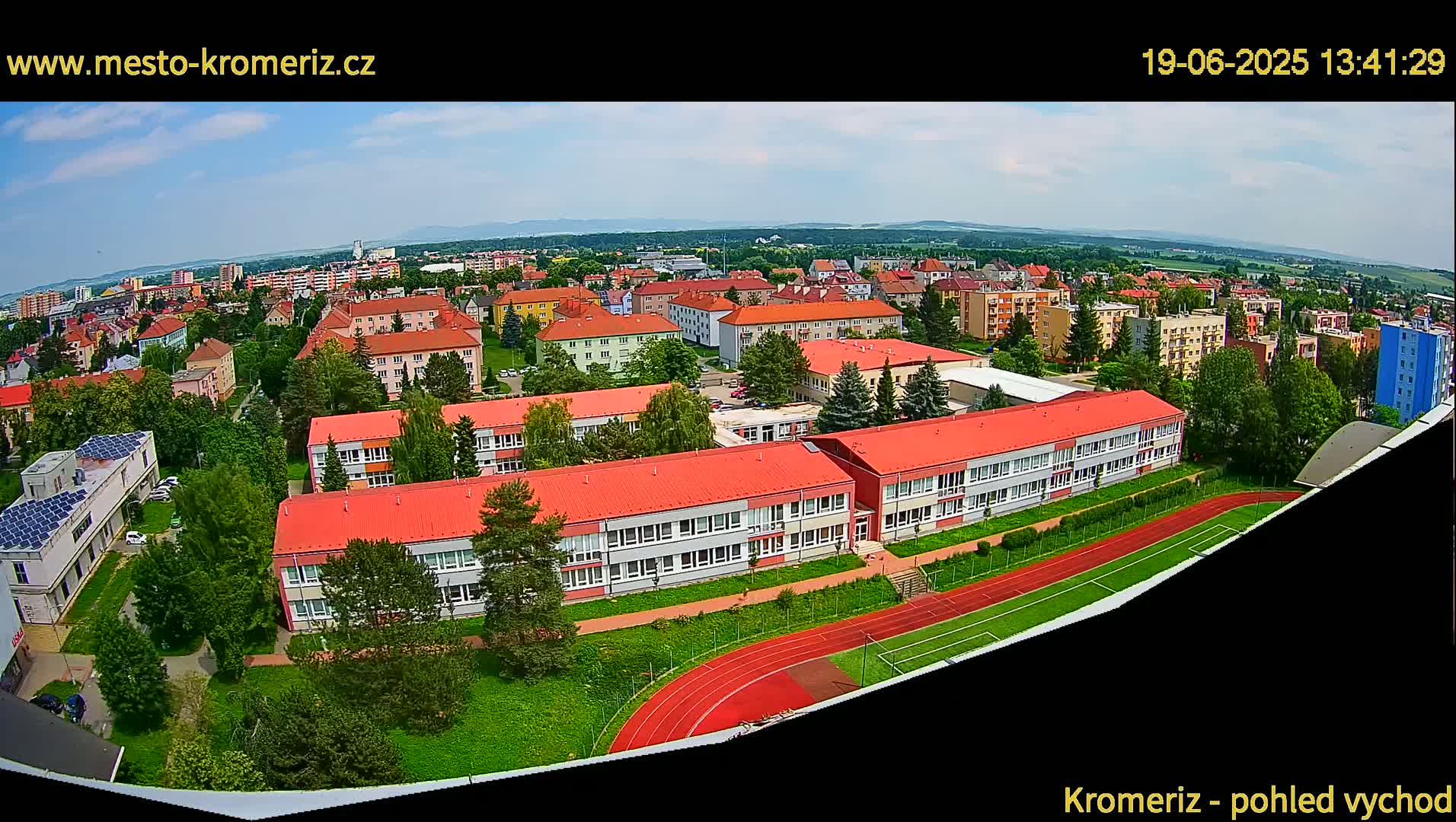 A high-angle view on a partly sunny day shows a large school complex with red roofs, surrounded by green landscaping, and a running track, situated within a residential area extending to a distant mountain range.
