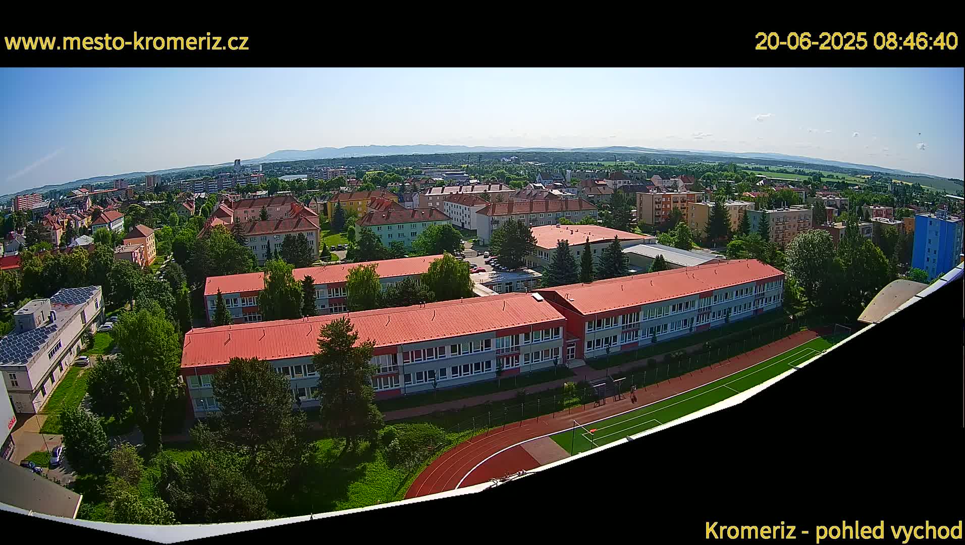 A wide aerial view on a sunny day shows a large school complex with red roofs and a running track, surrounded by other buildings and greenery, with distant mountains visible on the horizon.
