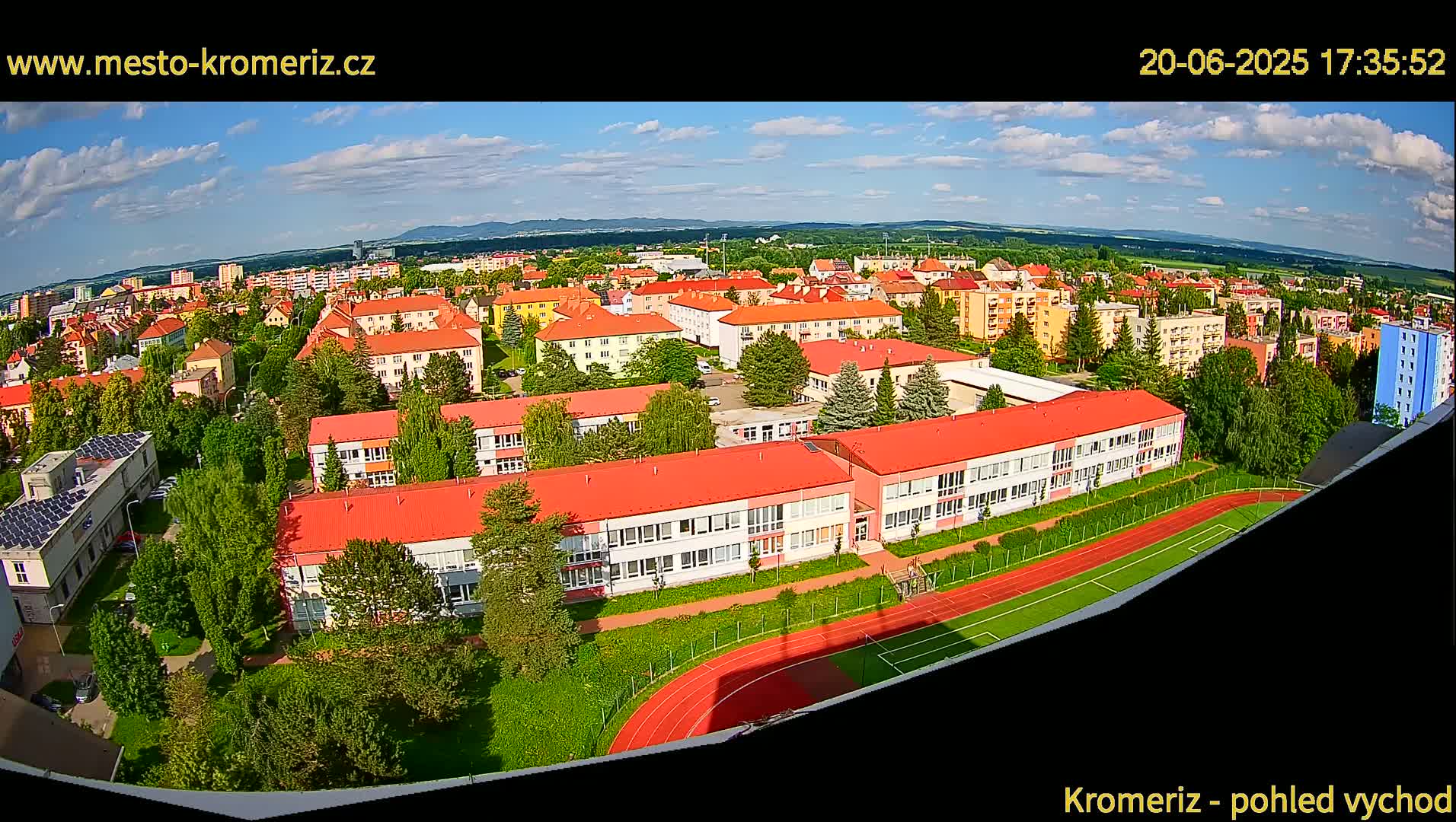 An aerial view of a town on a partly cloudy day shows a large school with a red roof and a running track next to it, surrounded by other buildings and green trees.