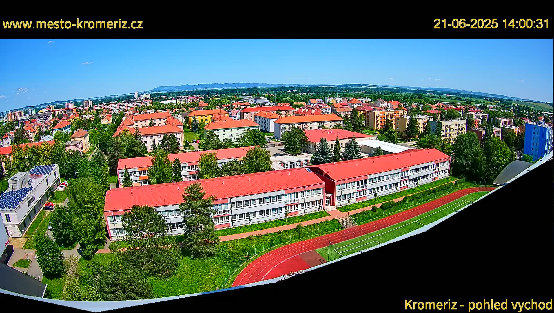 An aerial view on a sunny day shows a large school building with red roofs surrounded by other buildings and a running track.