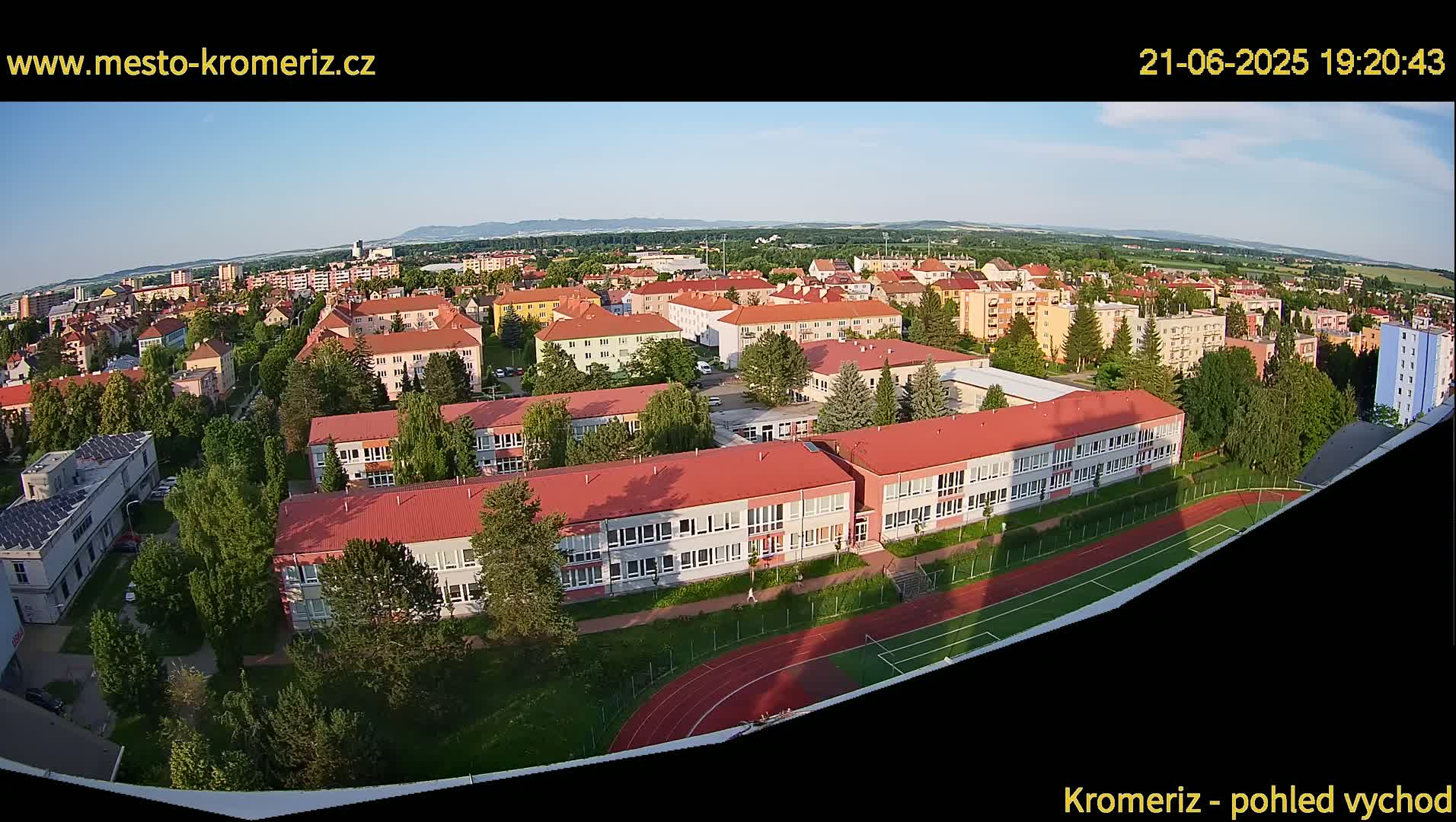 An aerial view shows a large school complex with red roofs and a running track, surrounded by residential buildings and trees under a clear evening sky.
