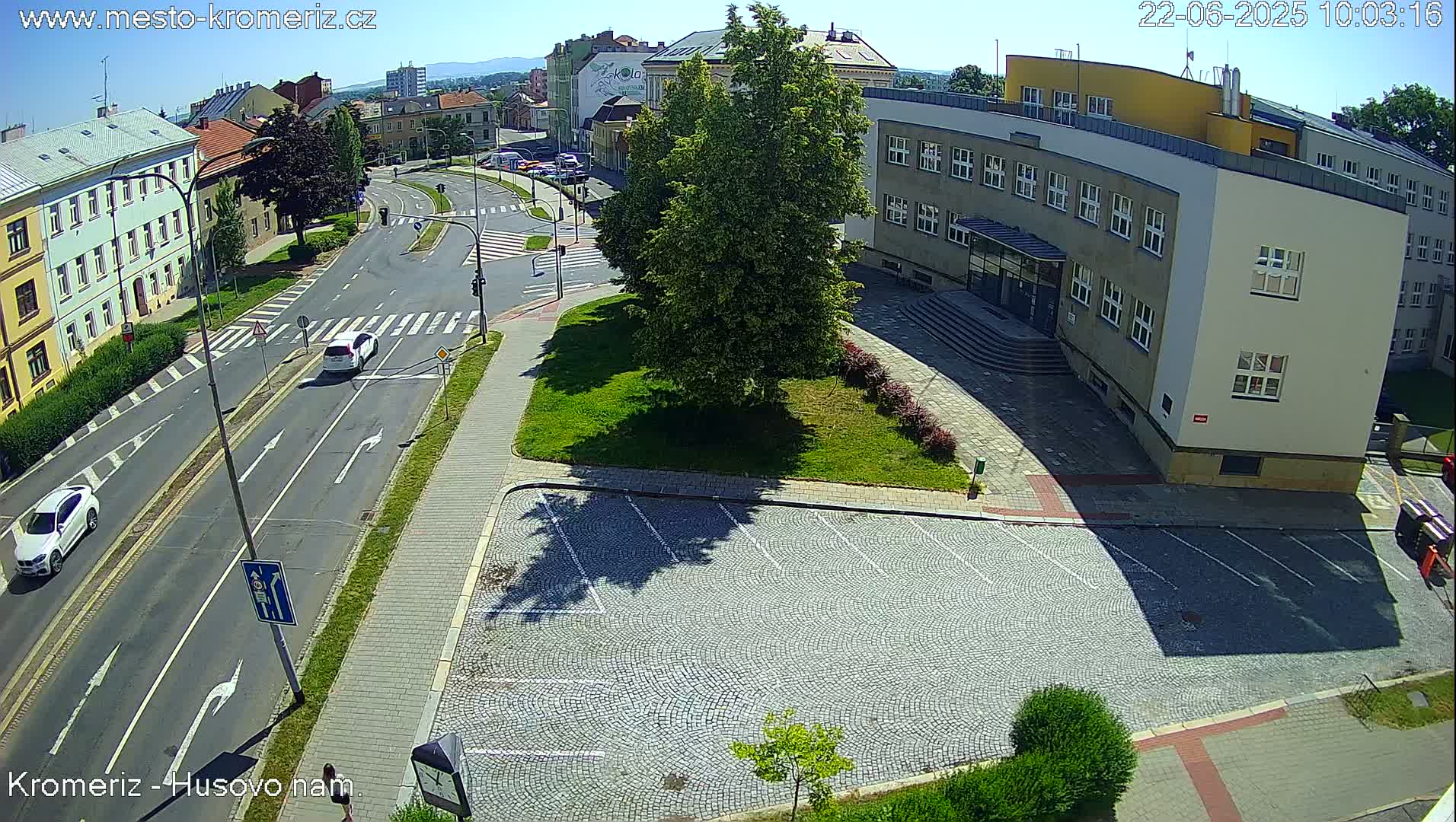 A sunny day reveals a street intersection with several cars, a large building with a parking lot, and green landscaping.