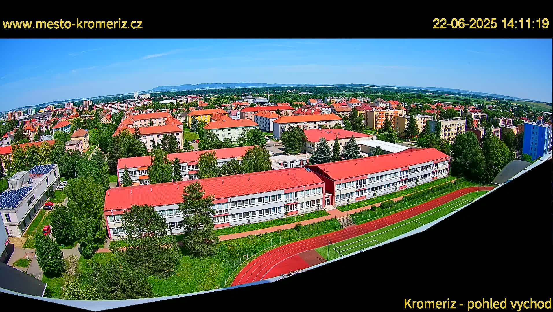 A wide aerial view shows a town on a sunny day, featuring a large school building with a red roof and a running track next to it, surrounded by other residential buildings and greenery.