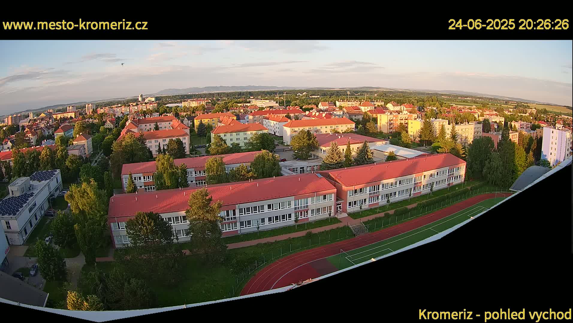An aerial view shows a large school complex with red roofs and a running track at sunset, surrounded by a residential area under a clear sky.