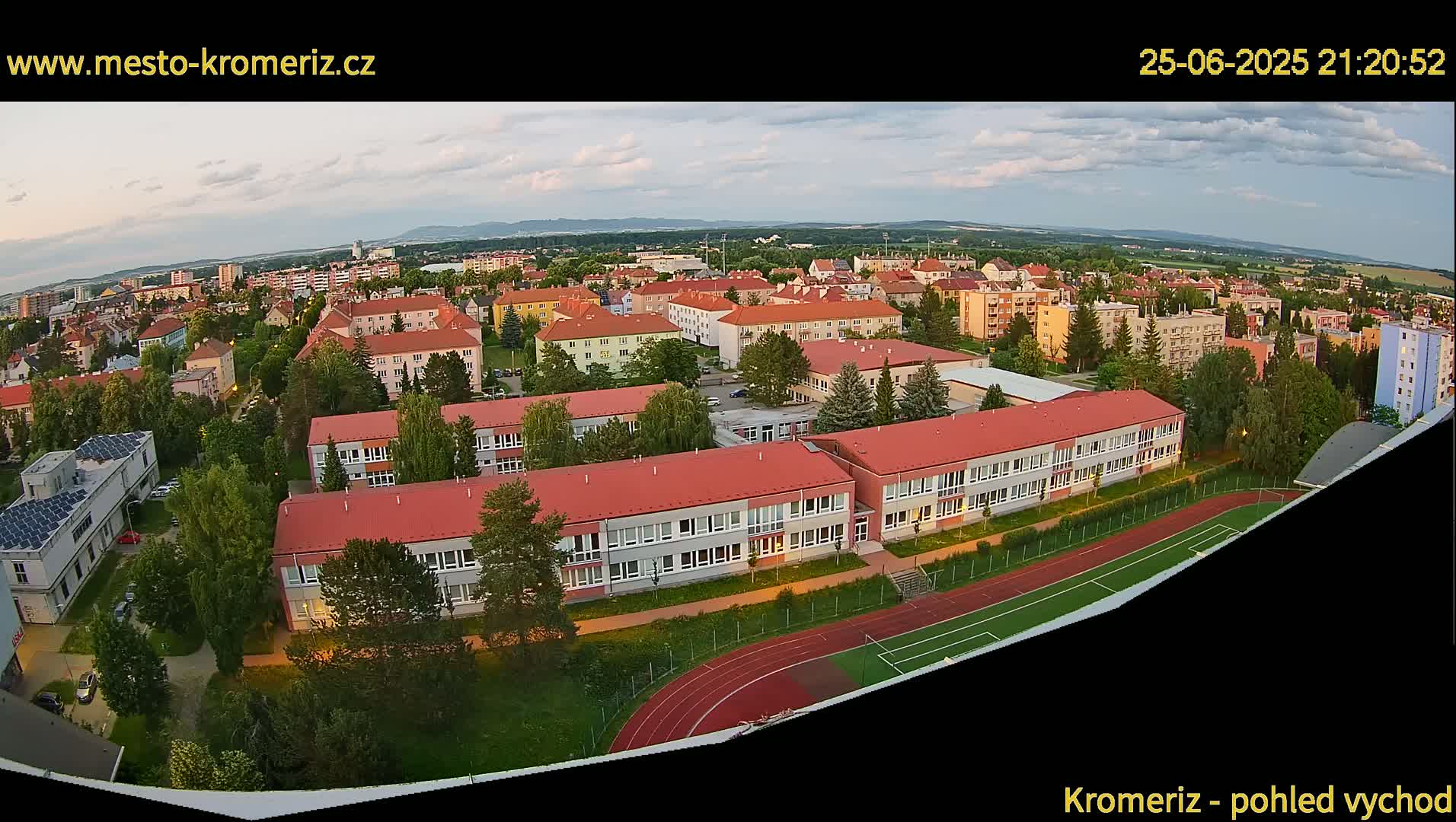 An aerial view at dusk shows a large school complex with red roofs and a running track, surrounded by residential buildings and trees under a partly cloudy sky.
