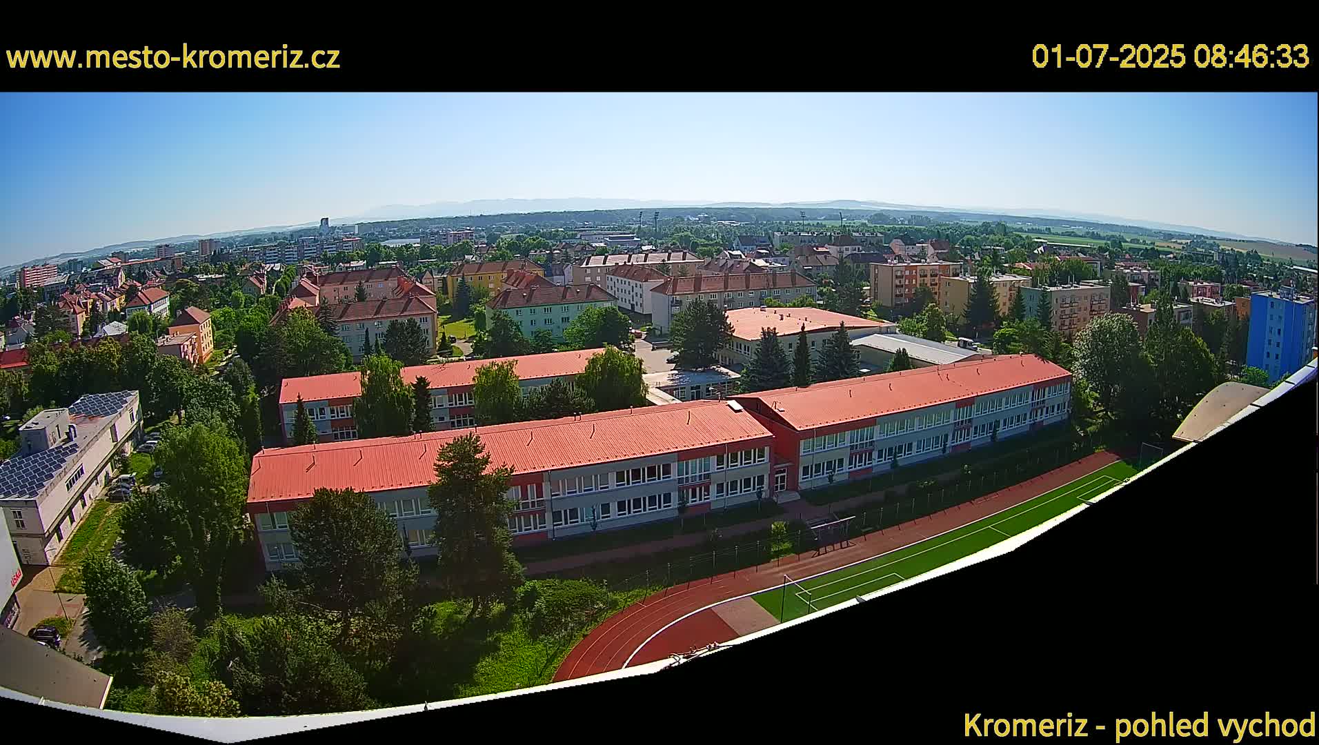 A high-angle, sunny view of a school and surrounding buildings, including a running track and soccer field, set within a residential area.