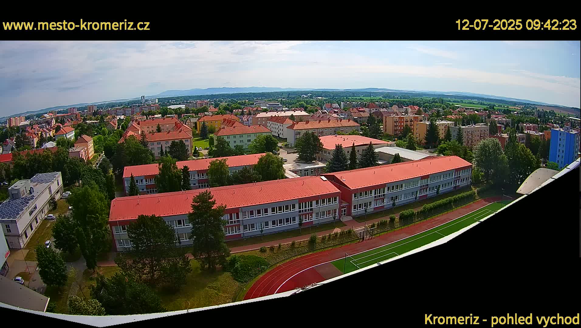 A high-angle, wide shot on a sunny day shows a large school complex with red roofs and a running track, surrounded by residential buildings and green trees, extending to distant mountains under a mostly clear sky.