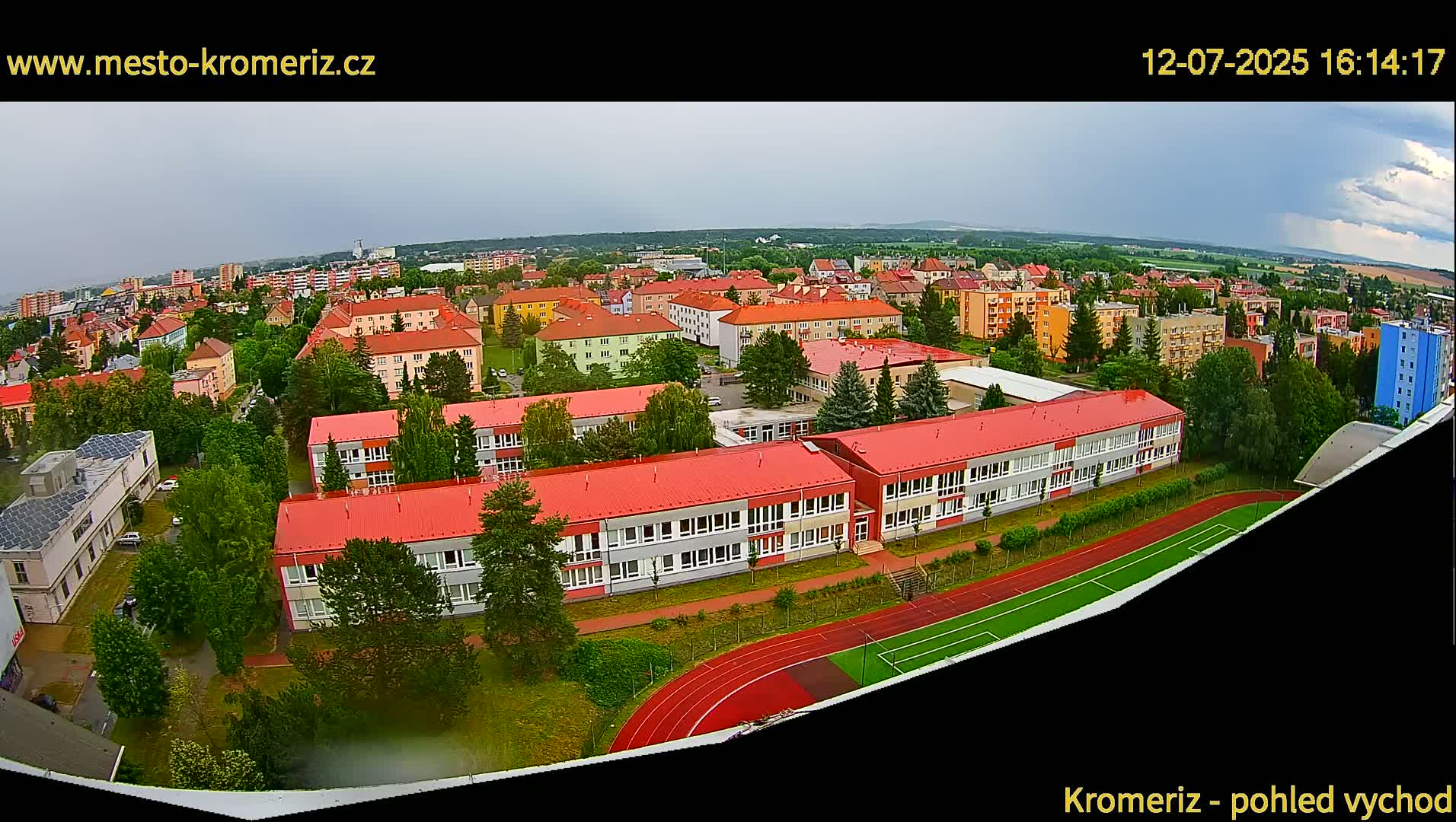 A high-angle, wide shot on a sunny day shows a large school complex with red roofs and a running track, surrounded by residential buildings and green trees, extending to distant mountains under a mostly clear sky.