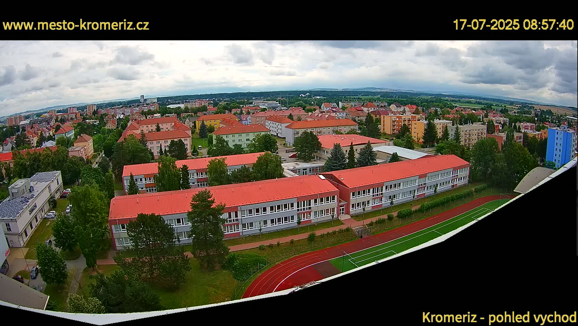An aerial view of a town on a partly cloudy day, showing numerous buildings, green spaces, and a running track.
