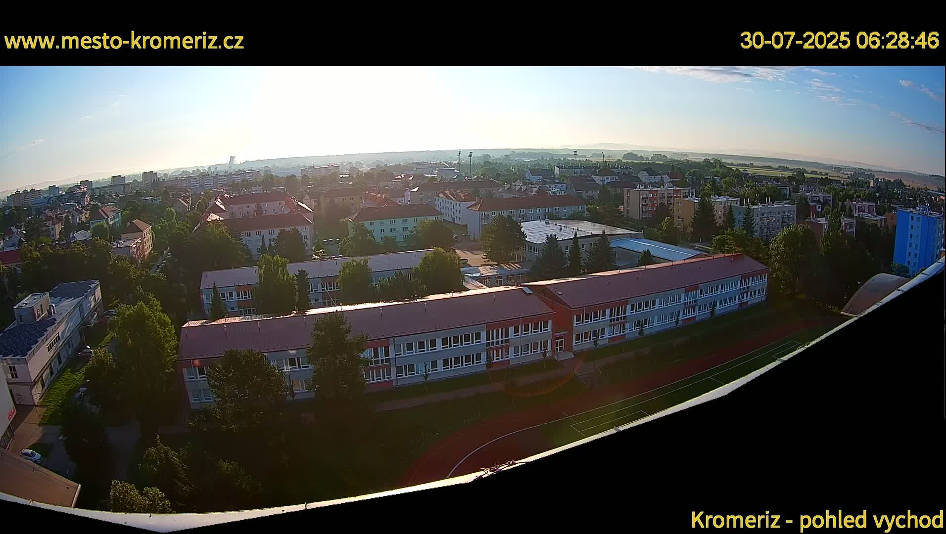 A high-angle, long shot on a clear day shows a large school building with a red roof, surrounded by other buildings and greenery, next to a running track and soccer field.
