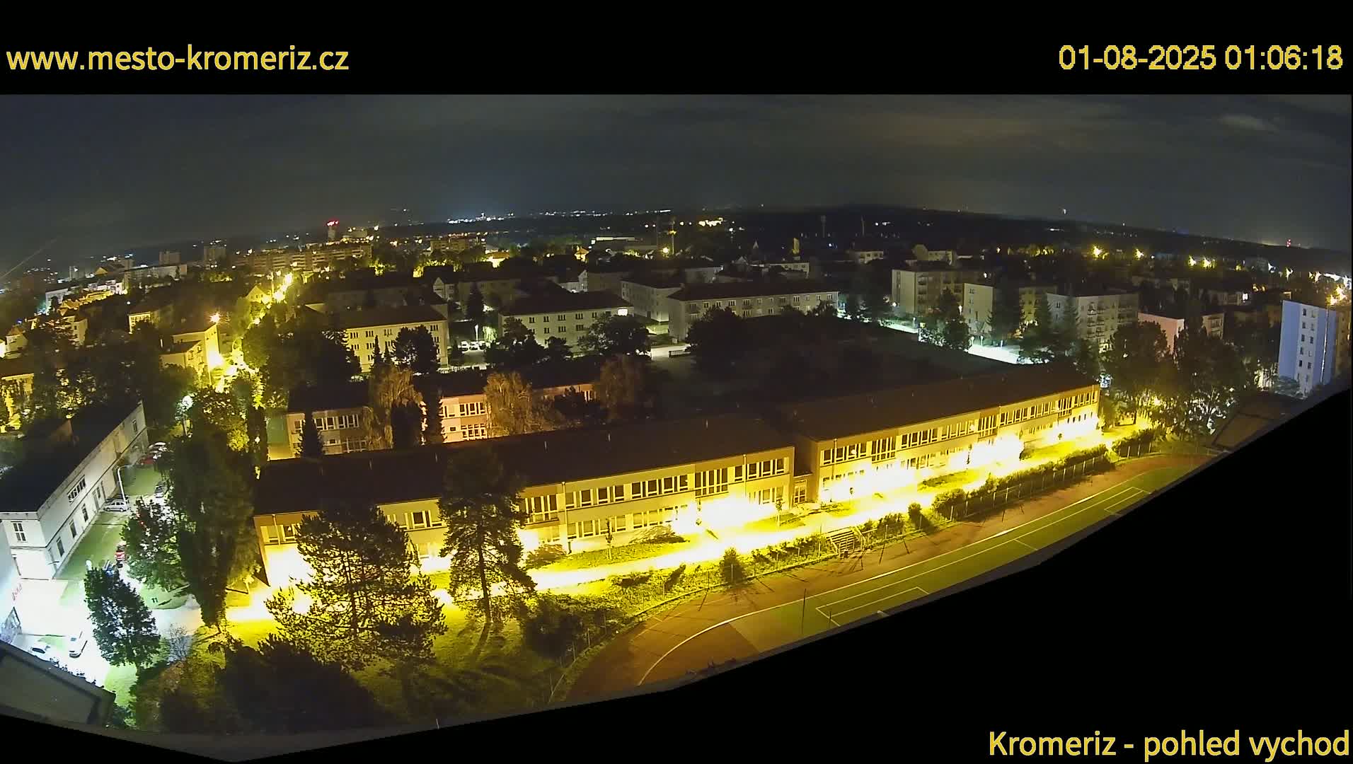 A nighttime aerial view shows a town with illuminated buildings and streets, a school with a sports field, and surrounding trees under a clear sky.