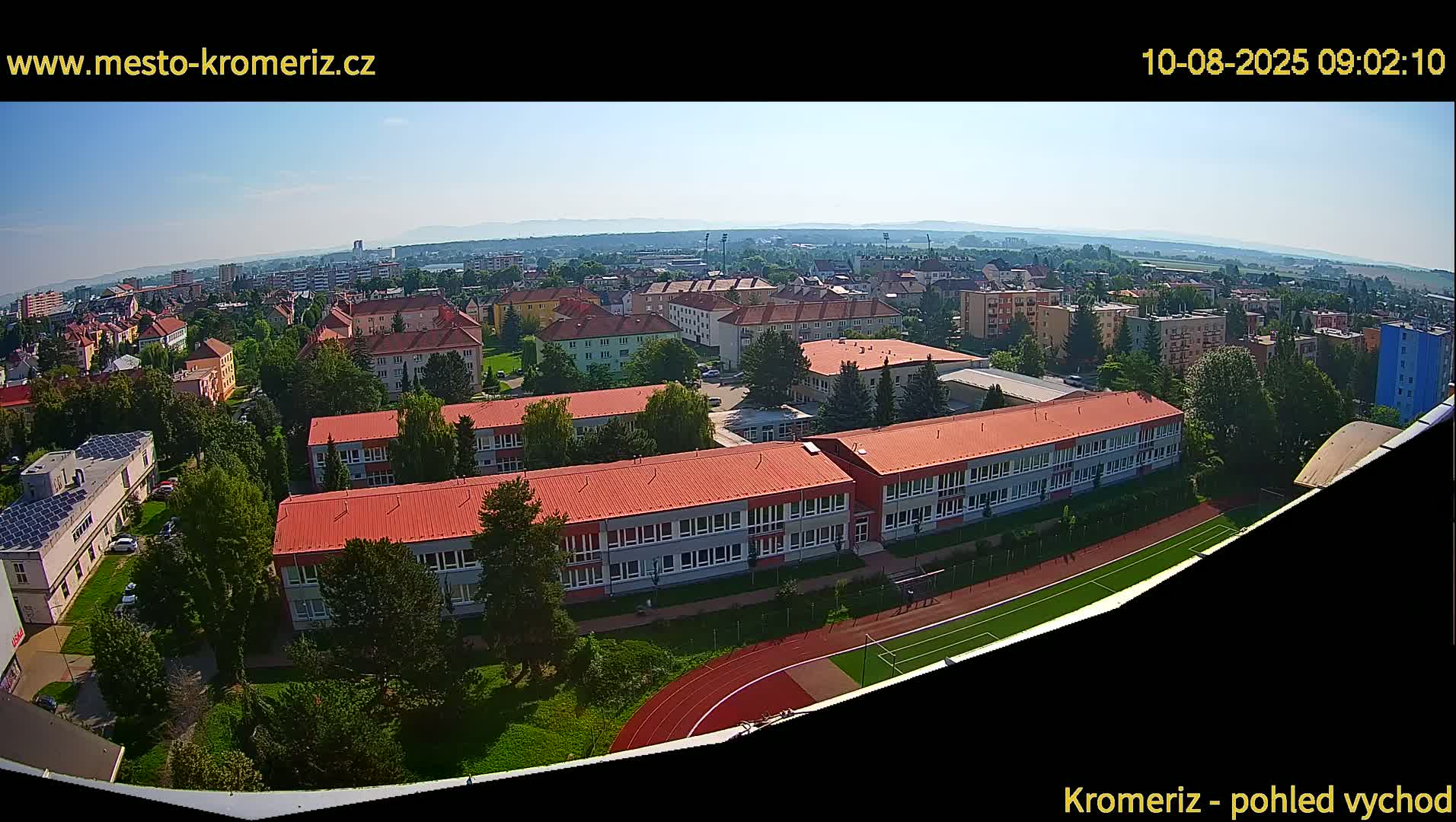 A high-angle, wide shot on a clear, sunny day shows a large school complex with red roofs, surrounded by other buildings and greenery, with a running track visible in the foreground.