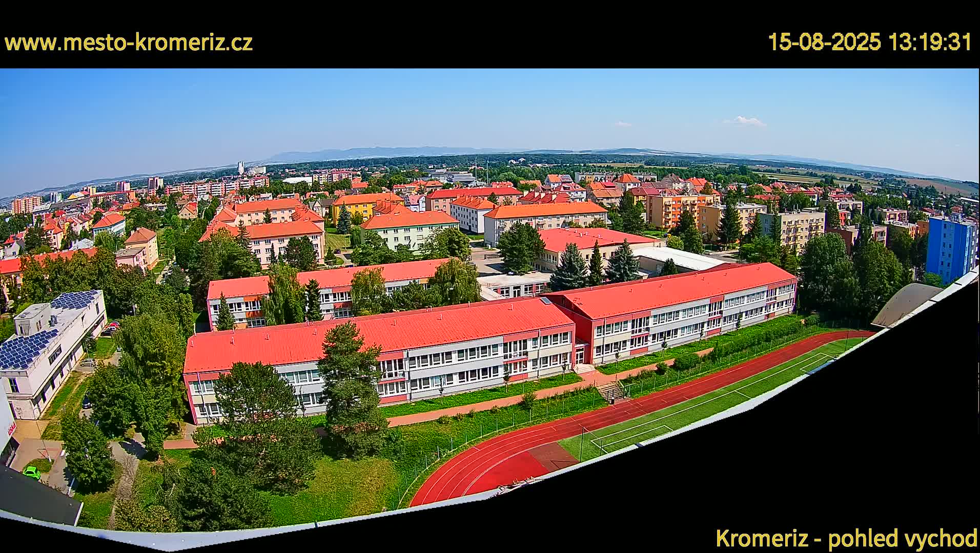 A high-angle, wide shot on a clear, sunny day shows a large school complex with red roofs, surrounded by other buildings and greenery, with a running track visible in the foreground.
