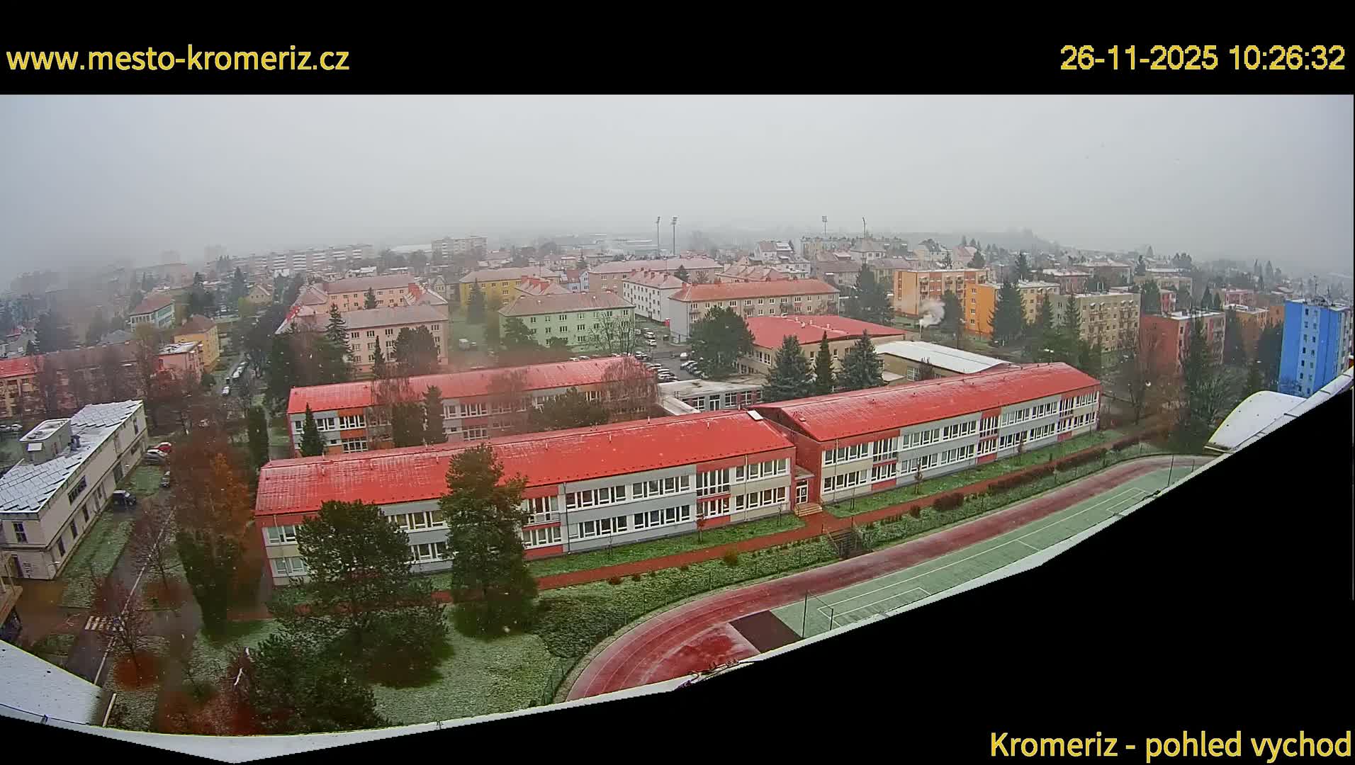A misty and snowy winter day view captures a townscape with several buildings featuring prominent red roofs and an athletic track in the foreground, backed by a sprawling residential area fading into the fog.