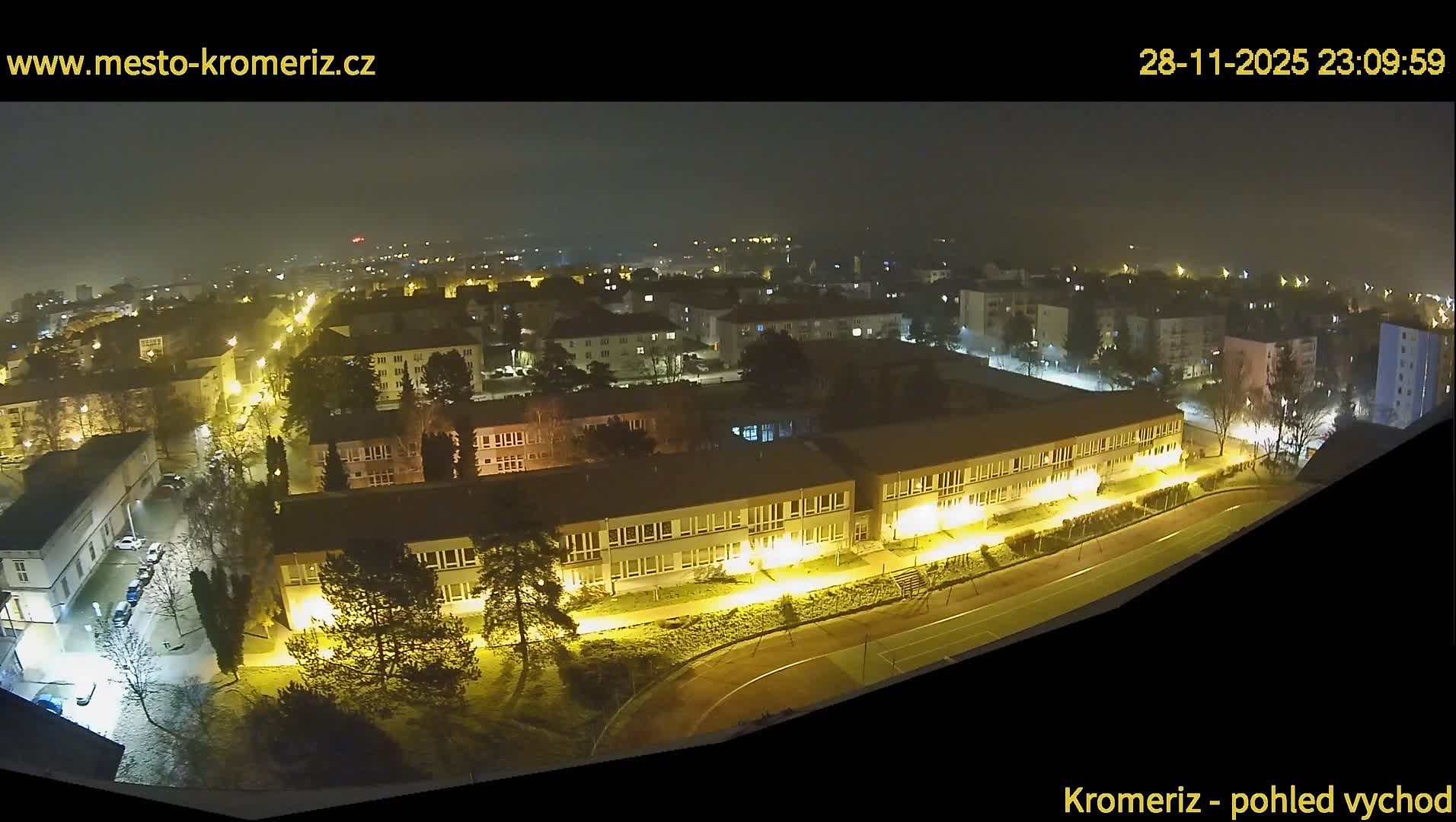 An elevated night view reveals a town's urban landscape under hazy conditions, with a brightly lit long building and an adjacent running track in the foreground, surrounded by other illuminated structures and trees.