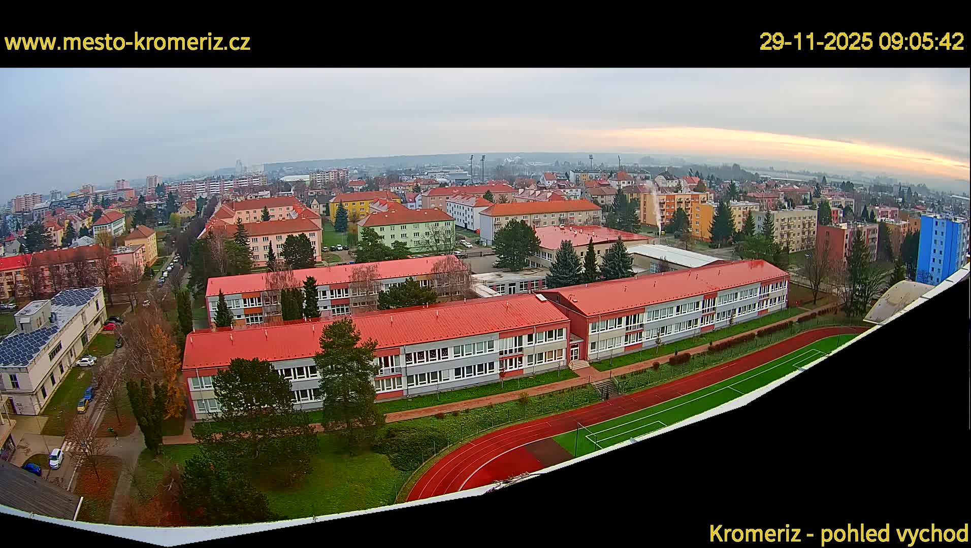 An overcast and hazy view captures an urban landscape featuring school buildings with red roofs, a track and field, and numerous residential structures extending into the distant, foggy horizon.