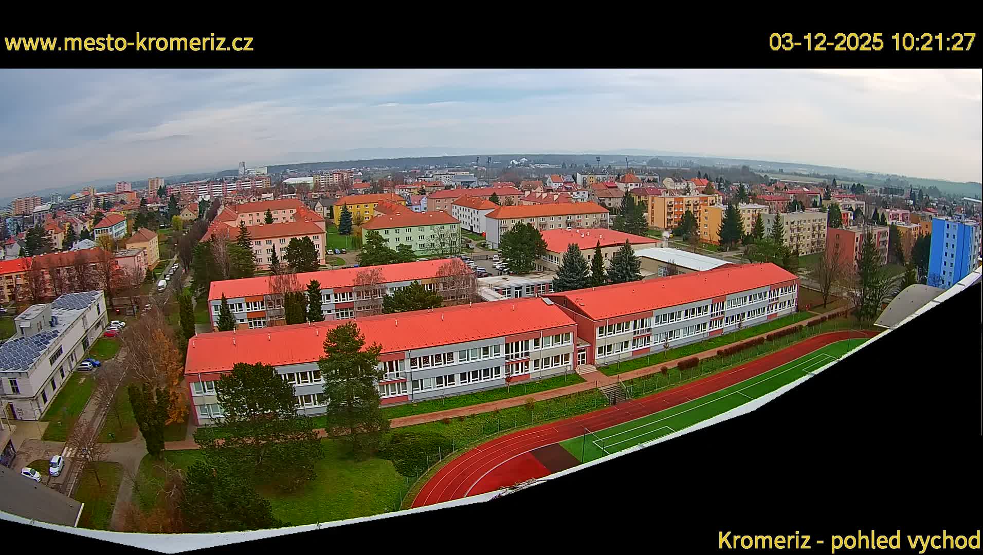Under a cloudy sky, an expansive daytime view reveals a town characterized by numerous red-roofed buildings, including a school complex with a running track and an adjacent sports field, amidst residential areas stretching towards distant hazy hills.