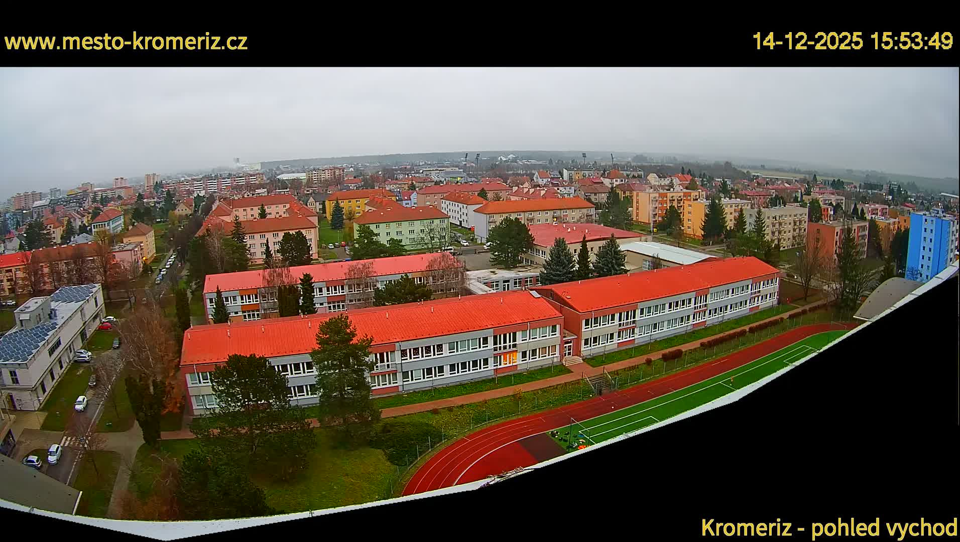 Under a cloudy sky, an expansive daytime view reveals a town characterized by numerous red-roofed buildings, including a school complex with a running track and an adjacent sports field, amidst residential areas stretching towards distant hazy hills.