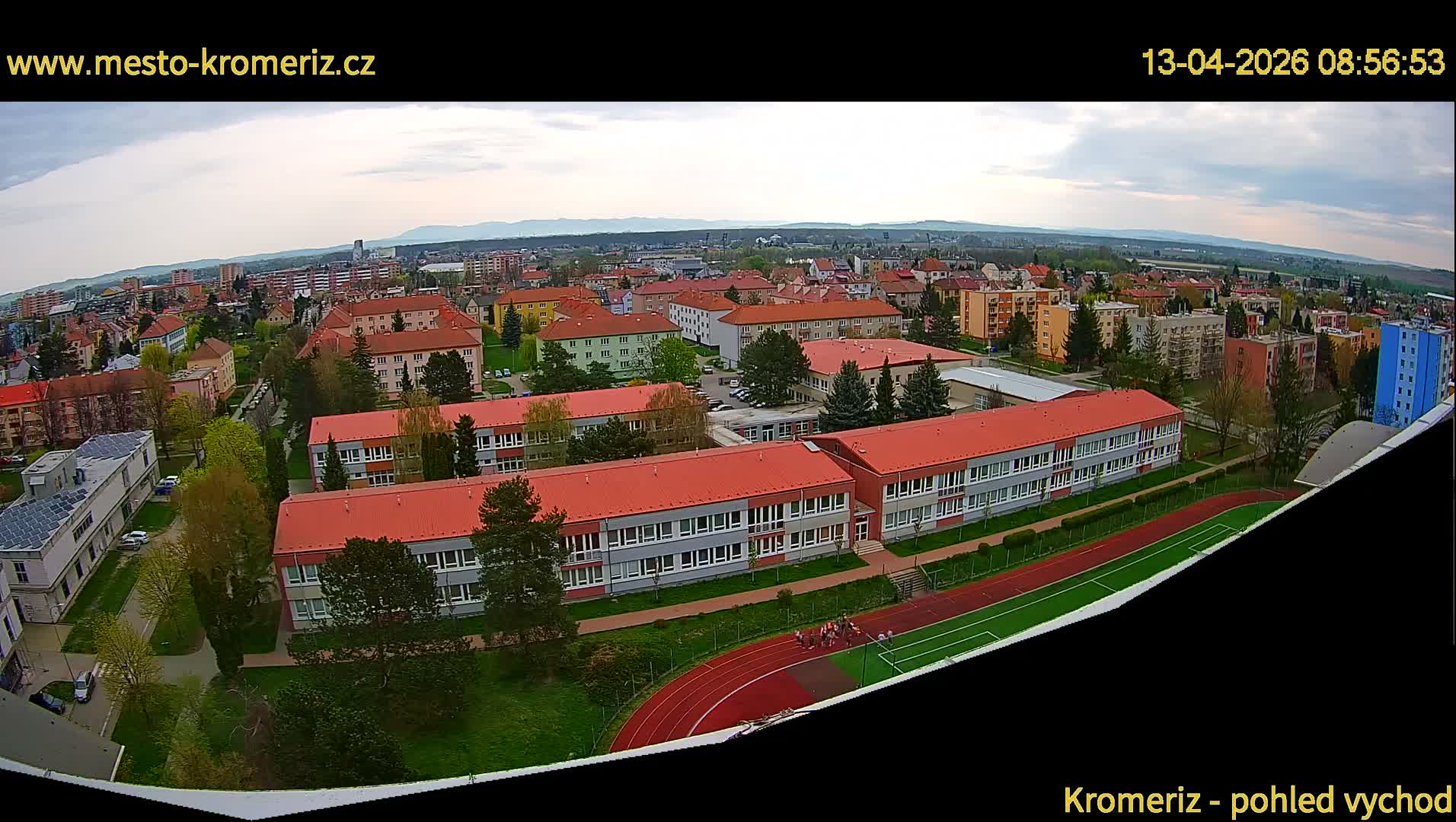 Under a cloudy sky, an expansive daytime view reveals a town characterized by numerous red-roofed buildings, including a school complex with a running track and an adjacent sports field, amidst residential areas stretching towards distant hazy hills.