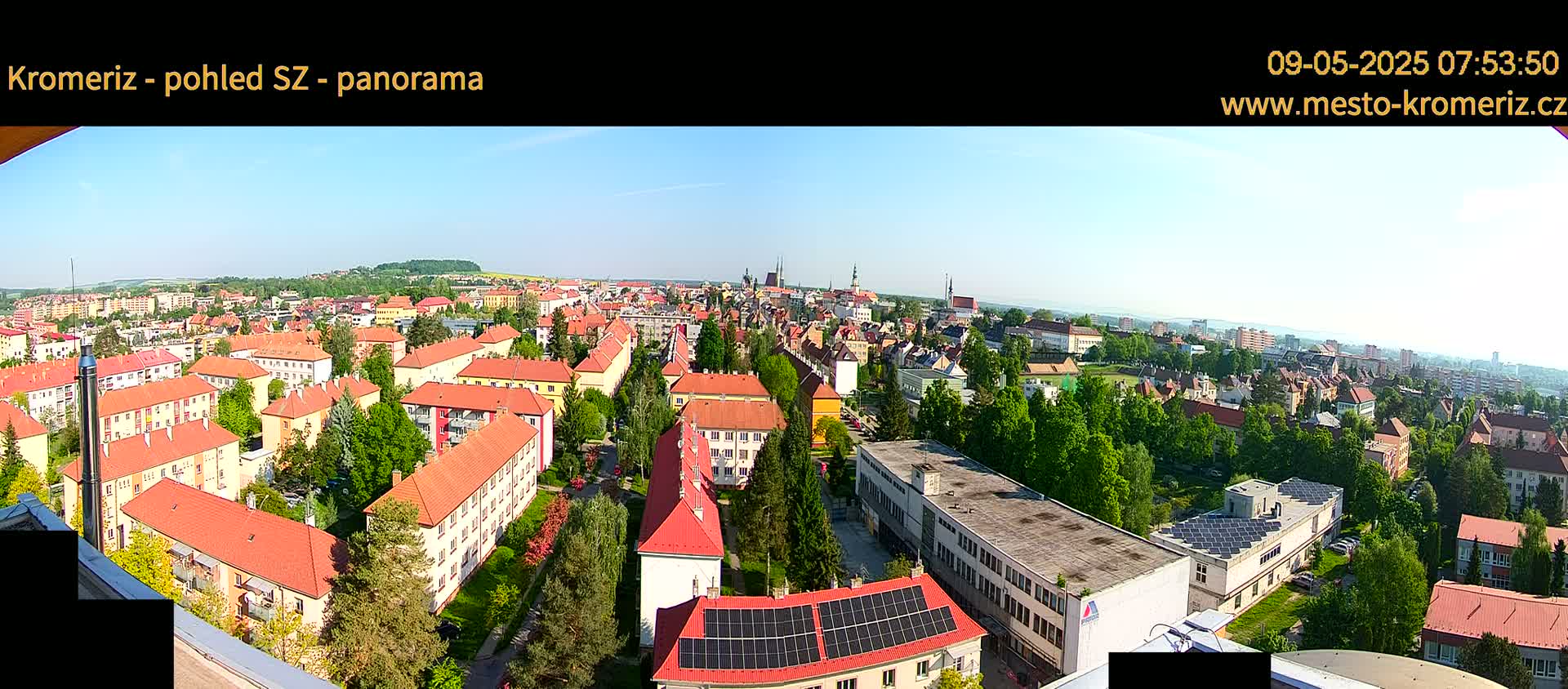A panoramic view of a town on a sunny day, showing numerous buildings with red-tiled roofs, surrounded by lush green trees.