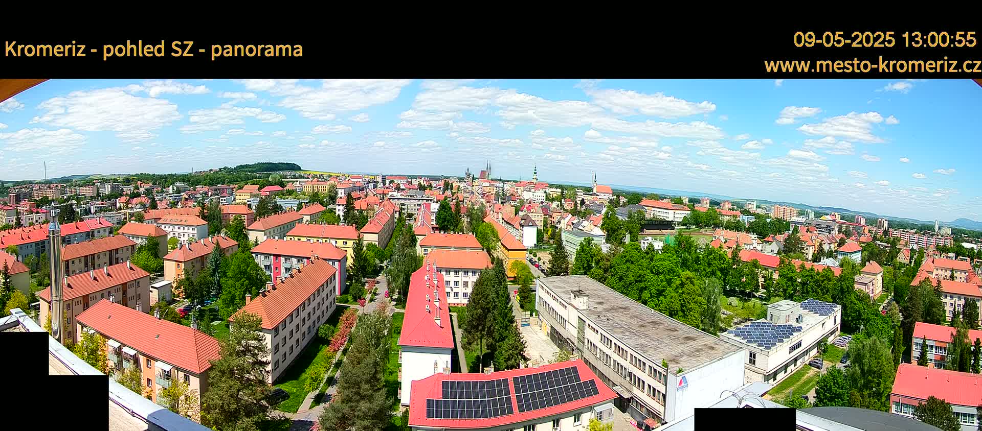 A panoramic view of a town on a partly sunny day, showing many red-roofed buildings and lush green trees.
