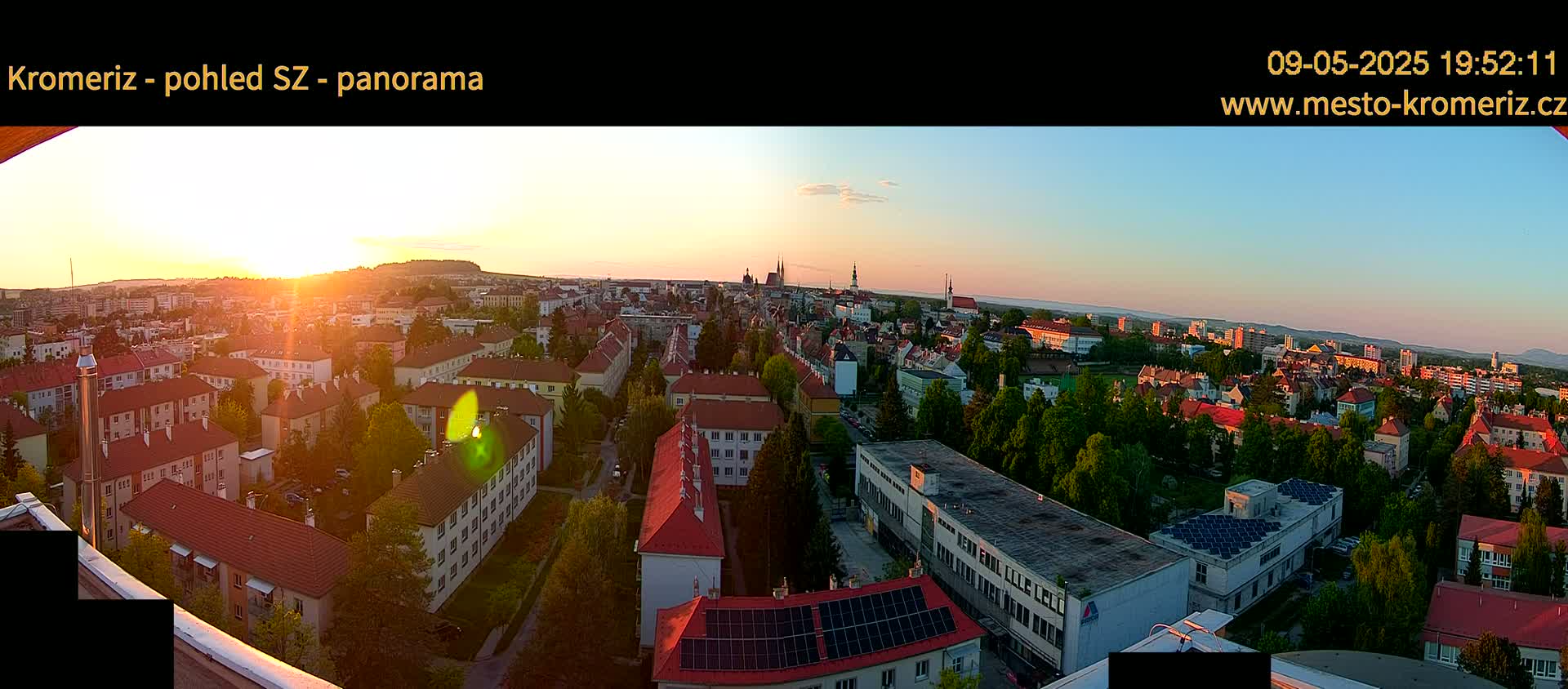 A panoramic view of a city at sunset, showing many buildings with red roofs and green trees, under a mostly clear sky.