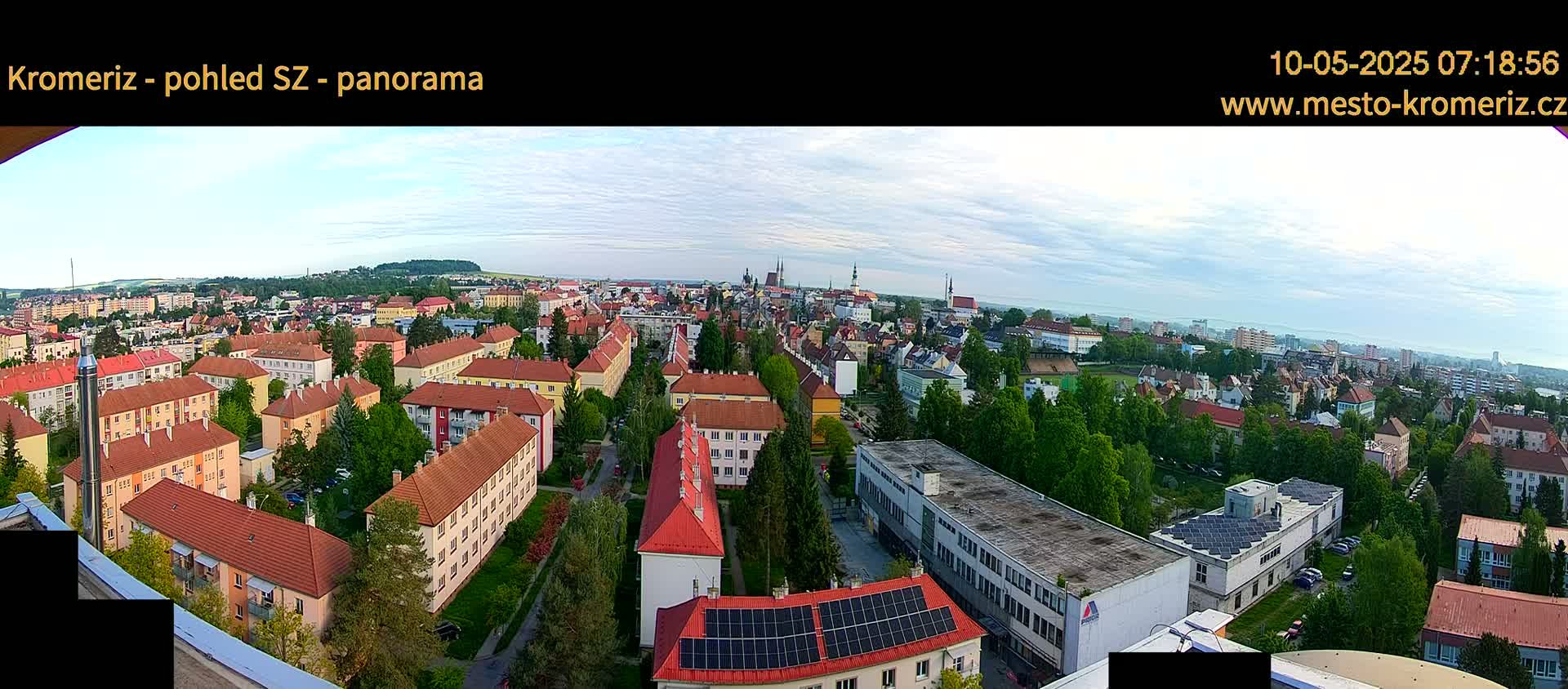 A panoramic view of a city on a partly cloudy day shows many low-rise buildings with red-tiled roofs, interspersed with green trees and a few taller buildings.
