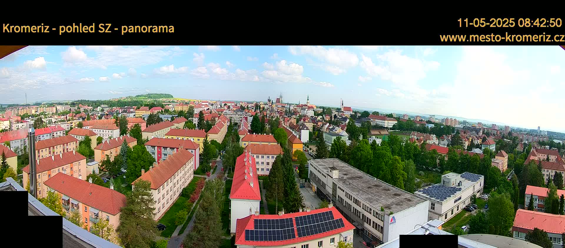 A panoramic view of a town on a partly cloudy day shows many low-rise buildings with red roofs, interspersed with green trees.