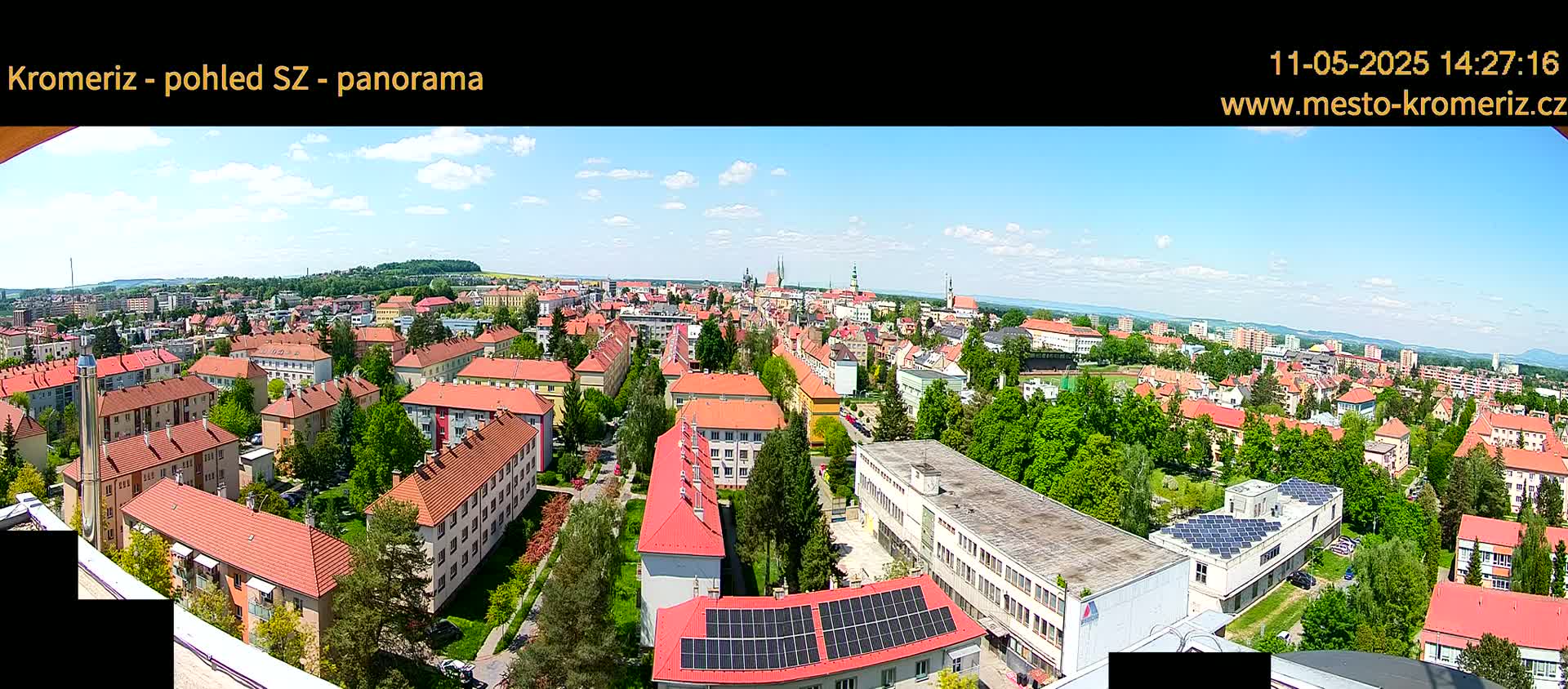 A panoramic view of a town on a partly sunny day, showing many buildings with red roofs and green trees interspersed among them.