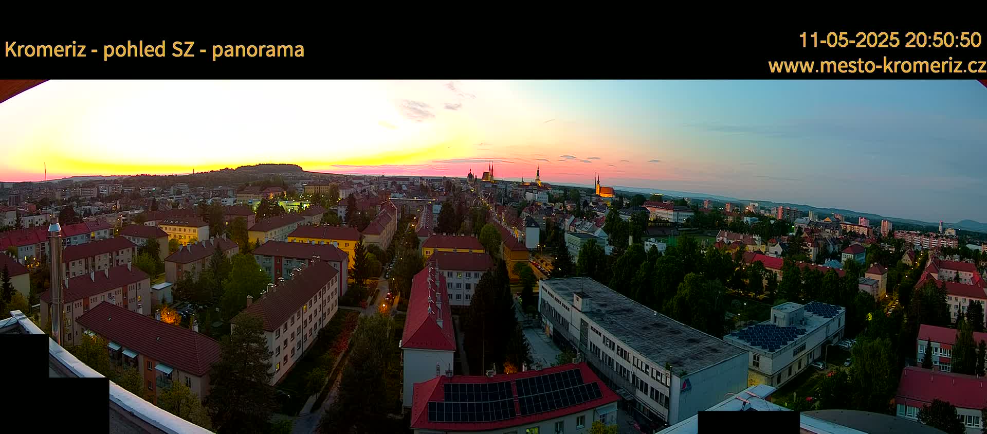 A panoramic view of a city at sunset, showing residential buildings and a church spire in the distance under a mostly clear sky.