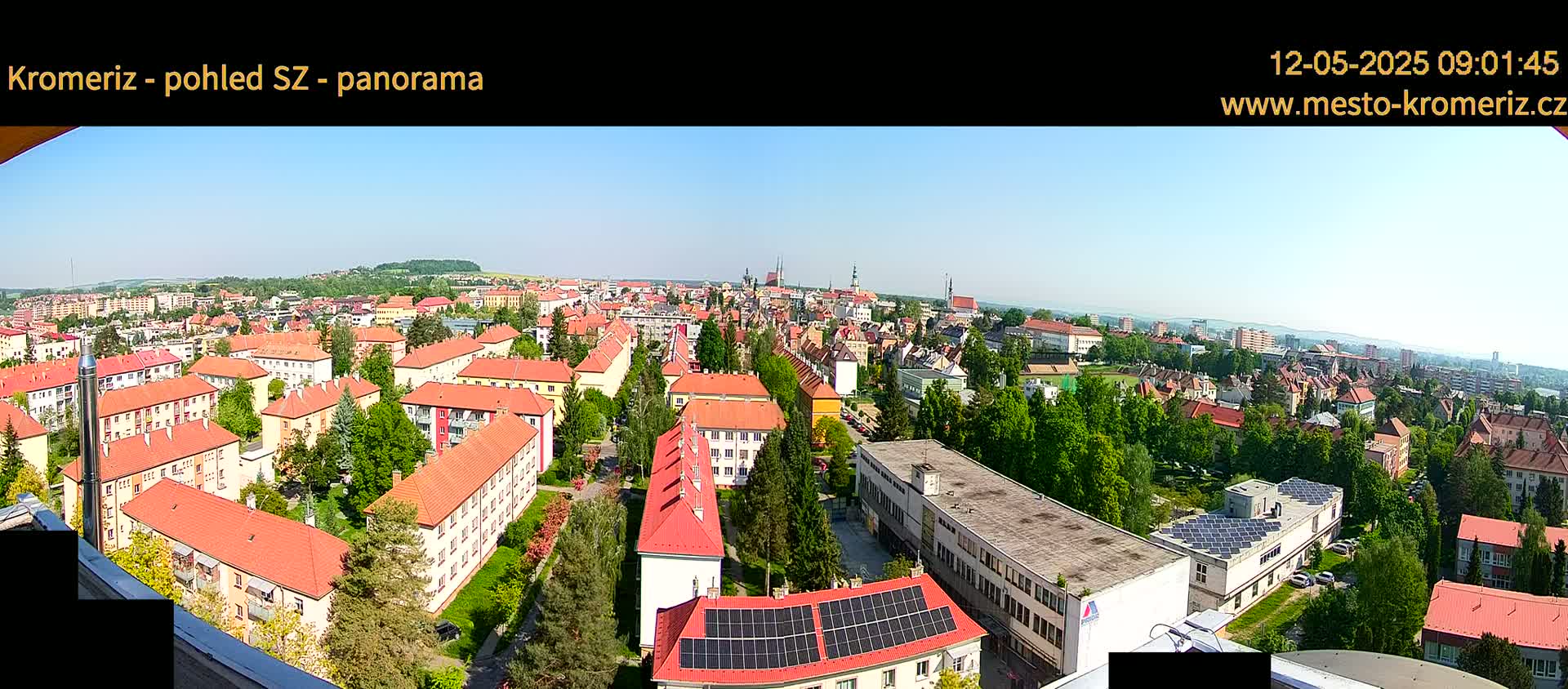 A panoramic view on a sunny day shows a sprawling town with many red-roofed buildings, interspersed with green trees and patches of grass, extending to a distant horizon.