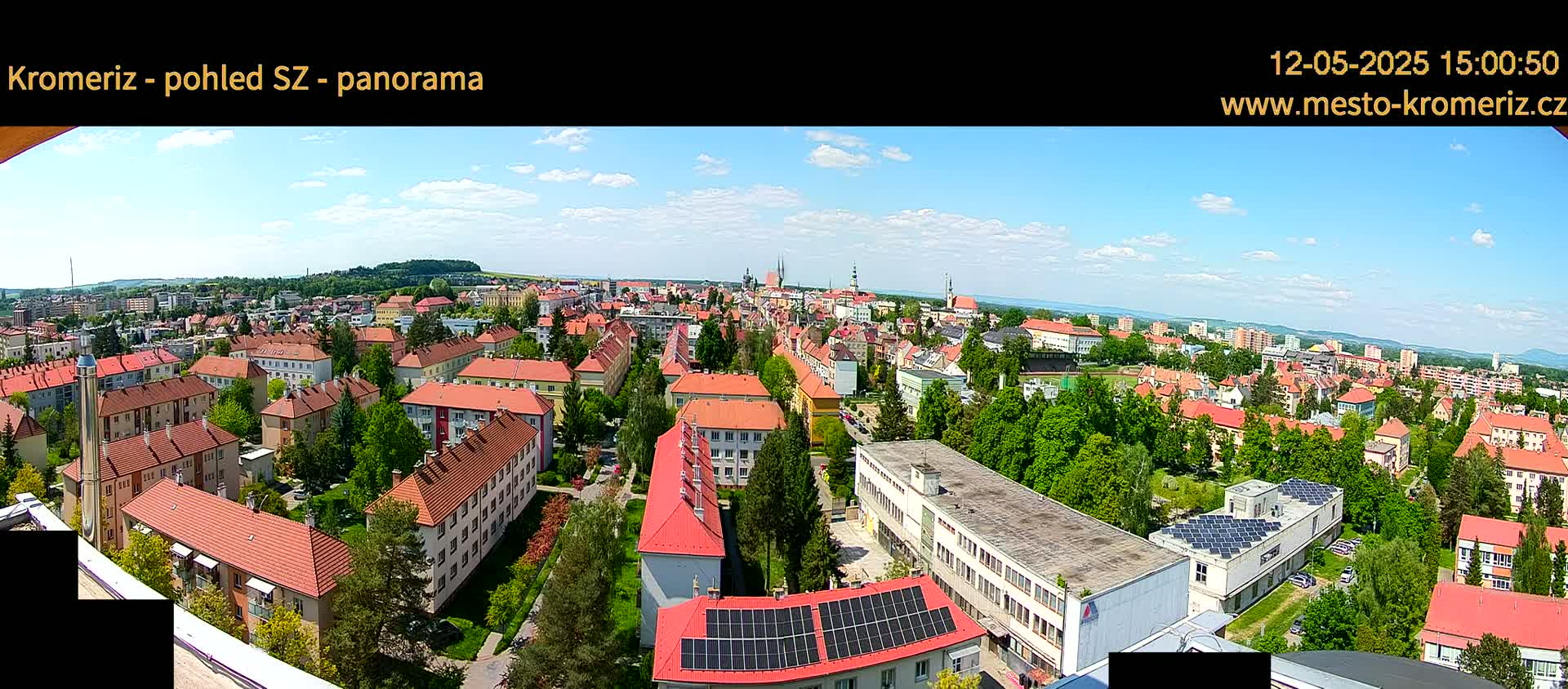 A panoramic view of a town on a partly sunny day, showing many buildings with red-tiled roofs and lush green trees.
