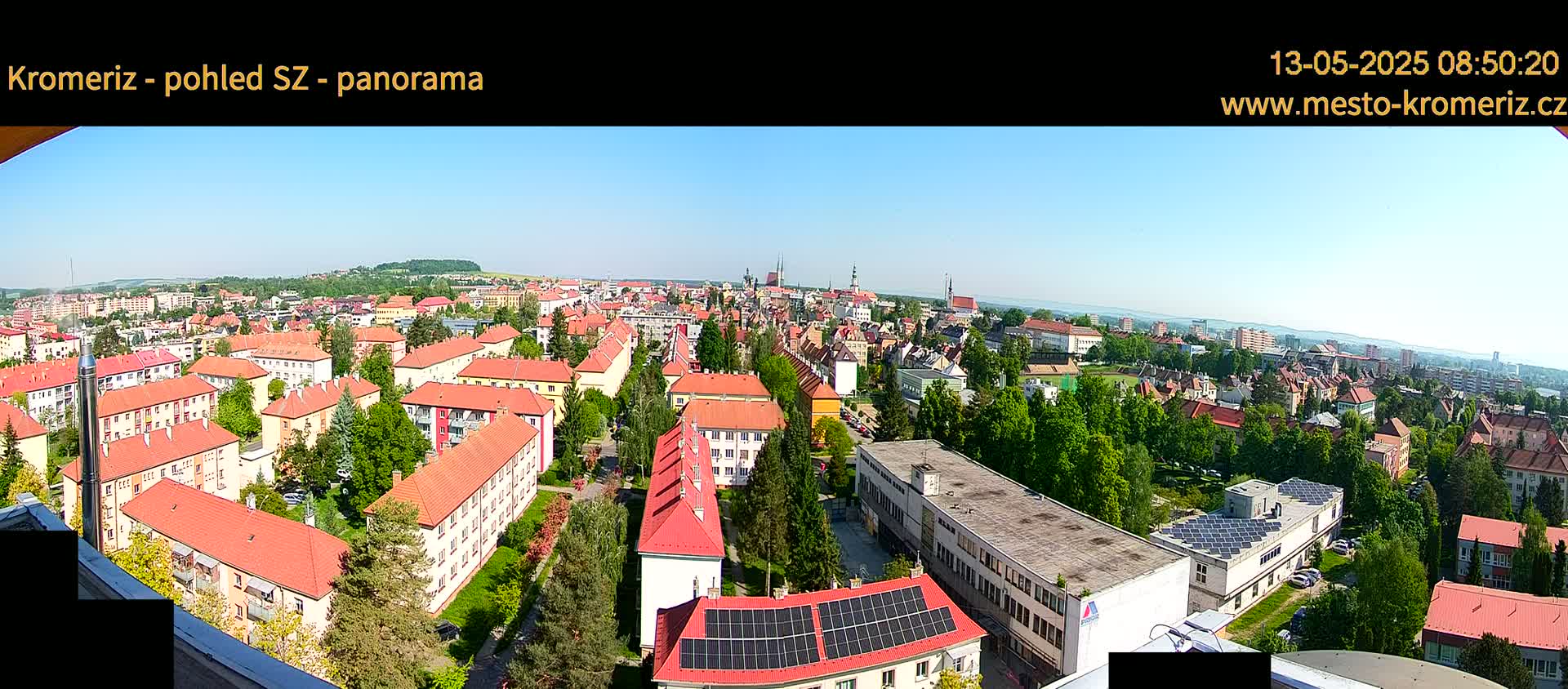 A panoramic view of a town on a clear, sunny day, showing numerous buildings with red-tiled roofs, interspersed with green trees and lawns.