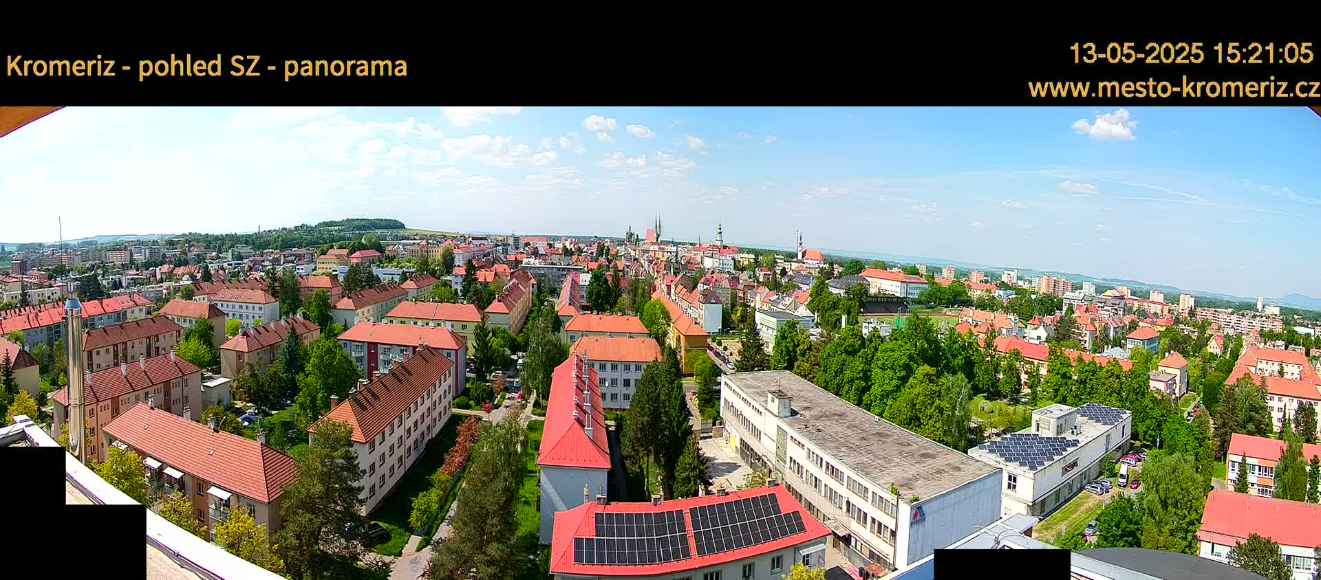 A panoramic view of a town under a mostly sunny sky shows numerous red-roofed buildings surrounded by lush green trees.
