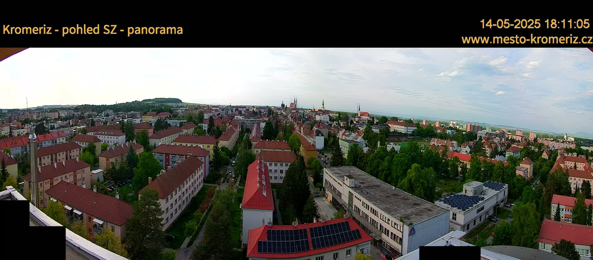 A panoramic view of a city with red-roofed buildings under a partly cloudy sky.