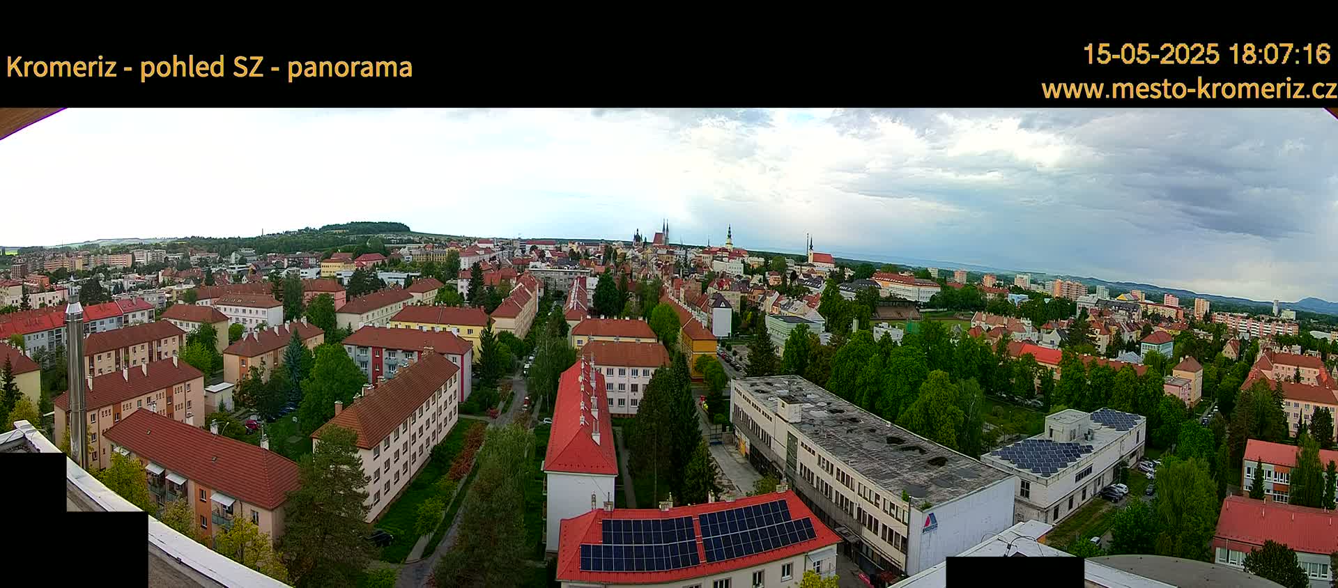 A panoramic view of a town on a partly cloudy day, showing numerous buildings, trees, and a distant horizon.