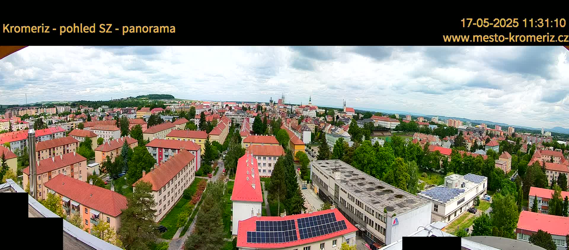 A panoramic view of a town on a partly cloudy day, showing numerous red-roofed buildings interspersed with green trees, with a larger building featuring solar panels in the foreground.