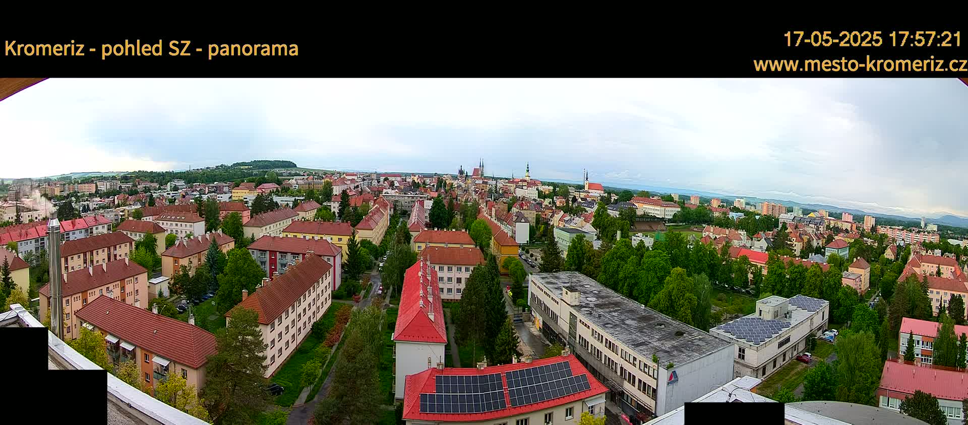 A panoramic view of a town on an overcast day, showing numerous residential buildings with red tile roofs, interspersed with green trees and a few larger buildings, including one with solar panels.