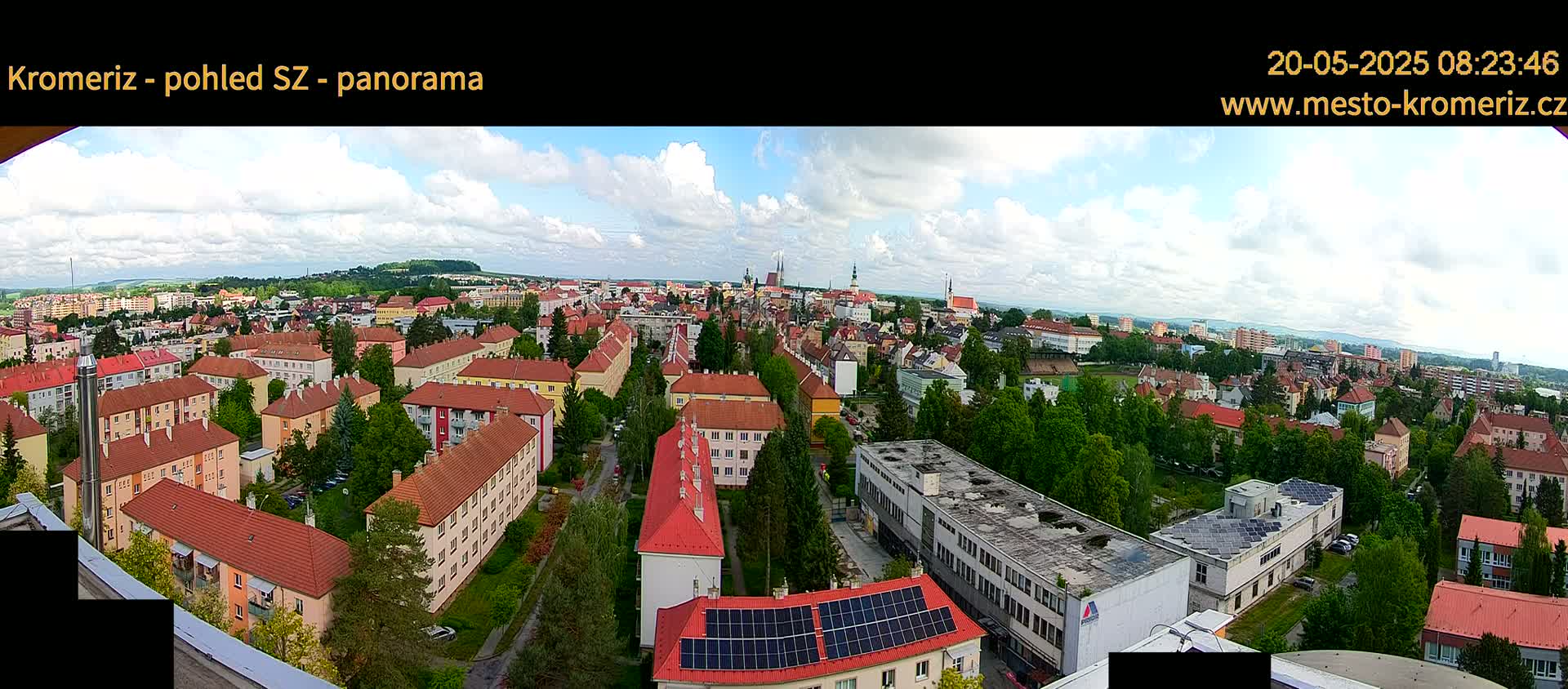 A panoramic view of a city under a partly cloudy sky shows many low-rise buildings with red roofs and green trees interspersed among them.