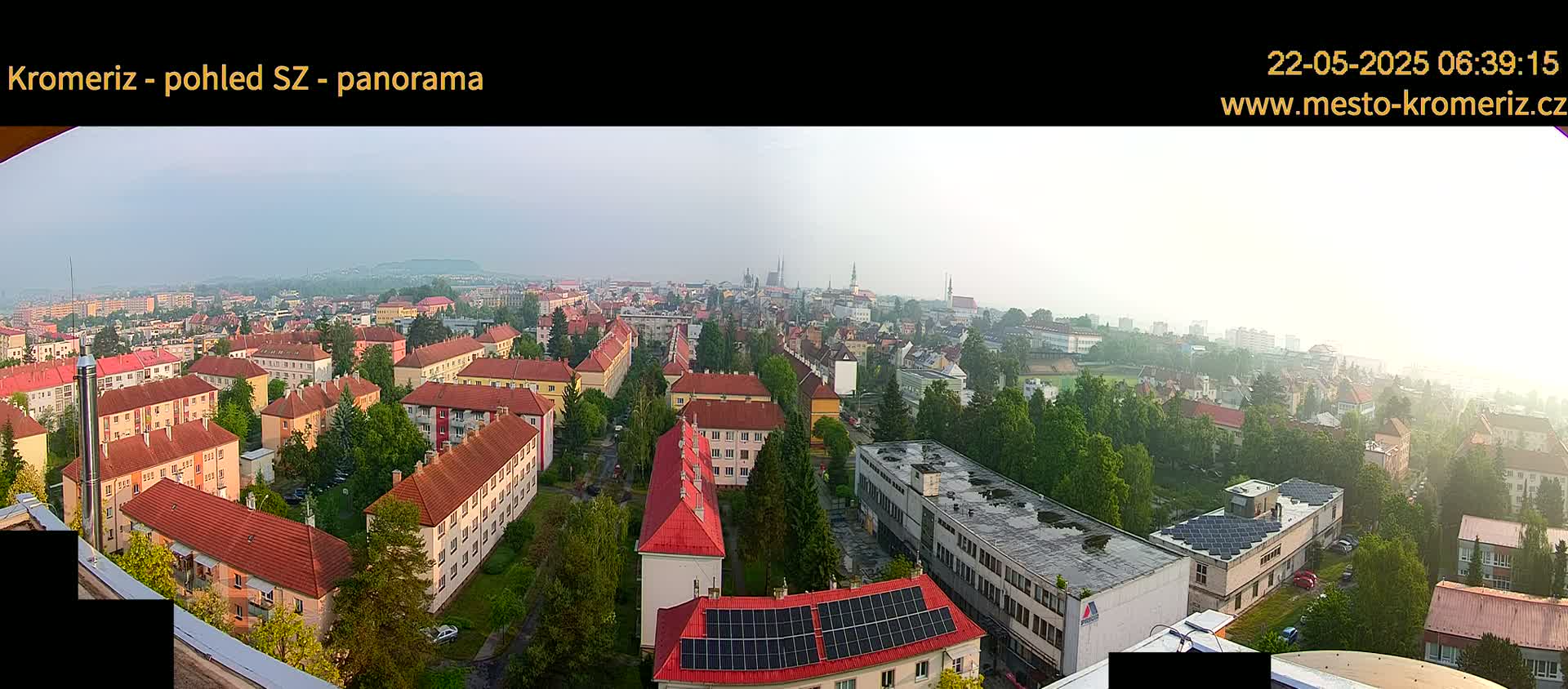 A hazy, panoramic view of a city with predominantly red-roofed buildings, interspersed with greenery, stretches to a distant horizon.