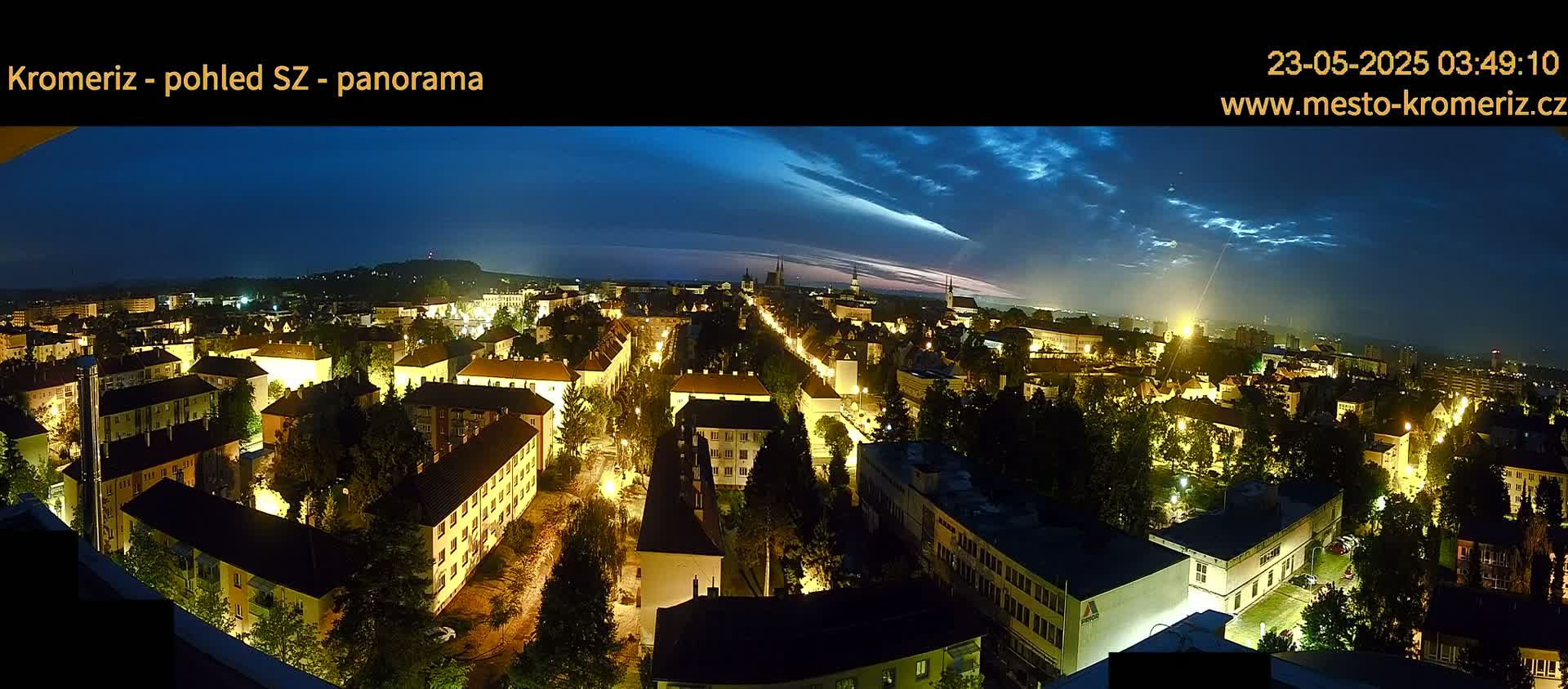 A nighttime panoramic view of a city with illuminated buildings and streets under a partly cloudy sky.