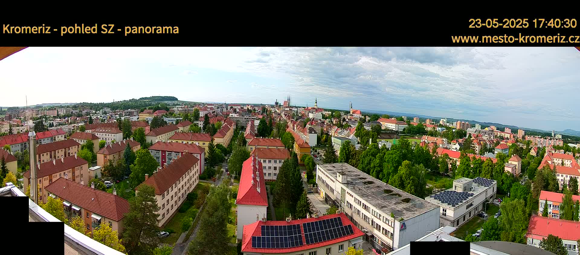 A panoramic view of a town with red-roofed buildings, green trees, and partly cloudy skies.