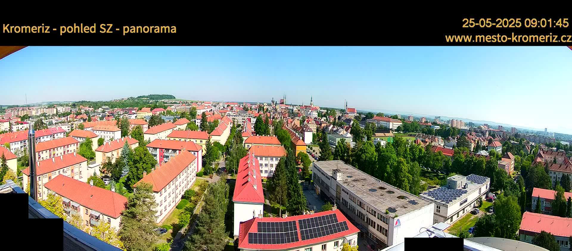 A panoramic view of a town on a clear, sunny day, showing many red-roofed buildings and green trees, with a distant horizon.