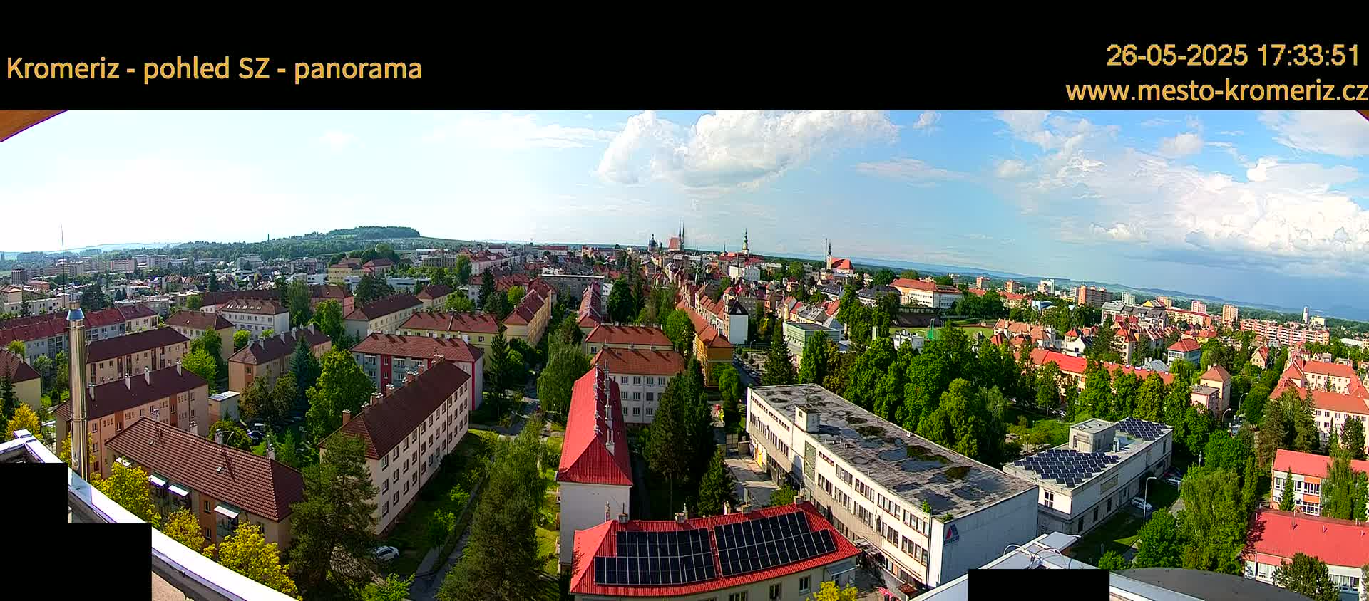 A panoramic view of a city on a partly sunny day, showing many buildings with red roofs, surrounded by green trees.