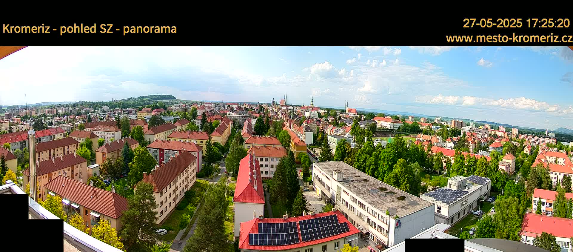 A panoramic view of a town on a partly cloudy day, showing many red-roofed buildings surrounded by green trees.
