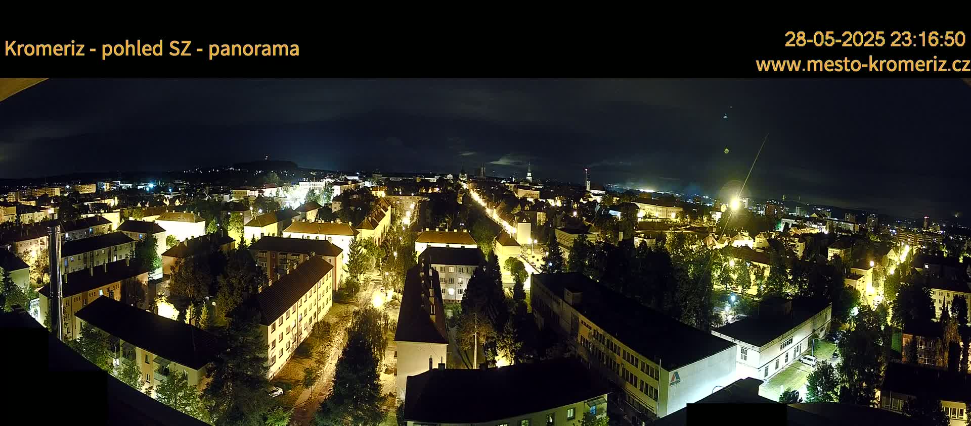 A nighttime panoramic view of a city with illuminated buildings and streets, surrounded by trees under a dark, mostly clear sky.