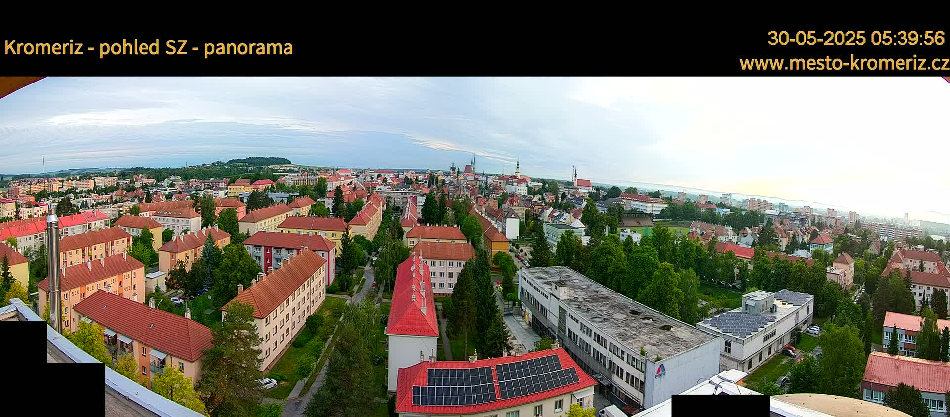 A panoramic view of a town, under a partly cloudy sky, shows numerous red-roofed buildings surrounded by green trees.