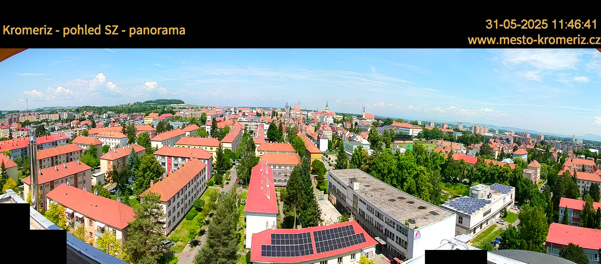 A panoramic view of a town on a sunny day with partly cloudy skies, showing many buildings with red-tiled roofs and green trees interspersed.