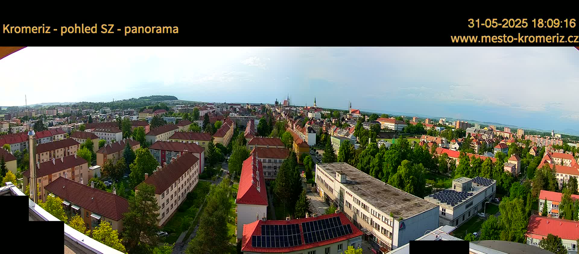 A panoramic view of a city on a partly cloudy day, showing numerous red-roofed buildings interspersed with green trees, extending to a horizon of distant hills.